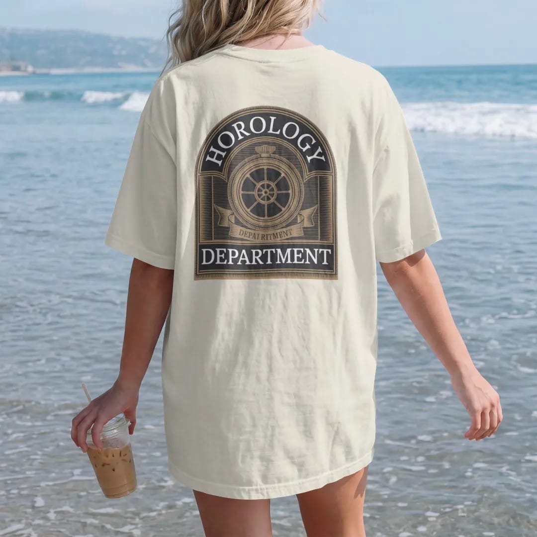 Woman on the beach in a Ivory tee, back view: a collegiate crest with a gear and balance wheel mascot under arched Horology and Department, as a fine engraving in antique gold and cream.