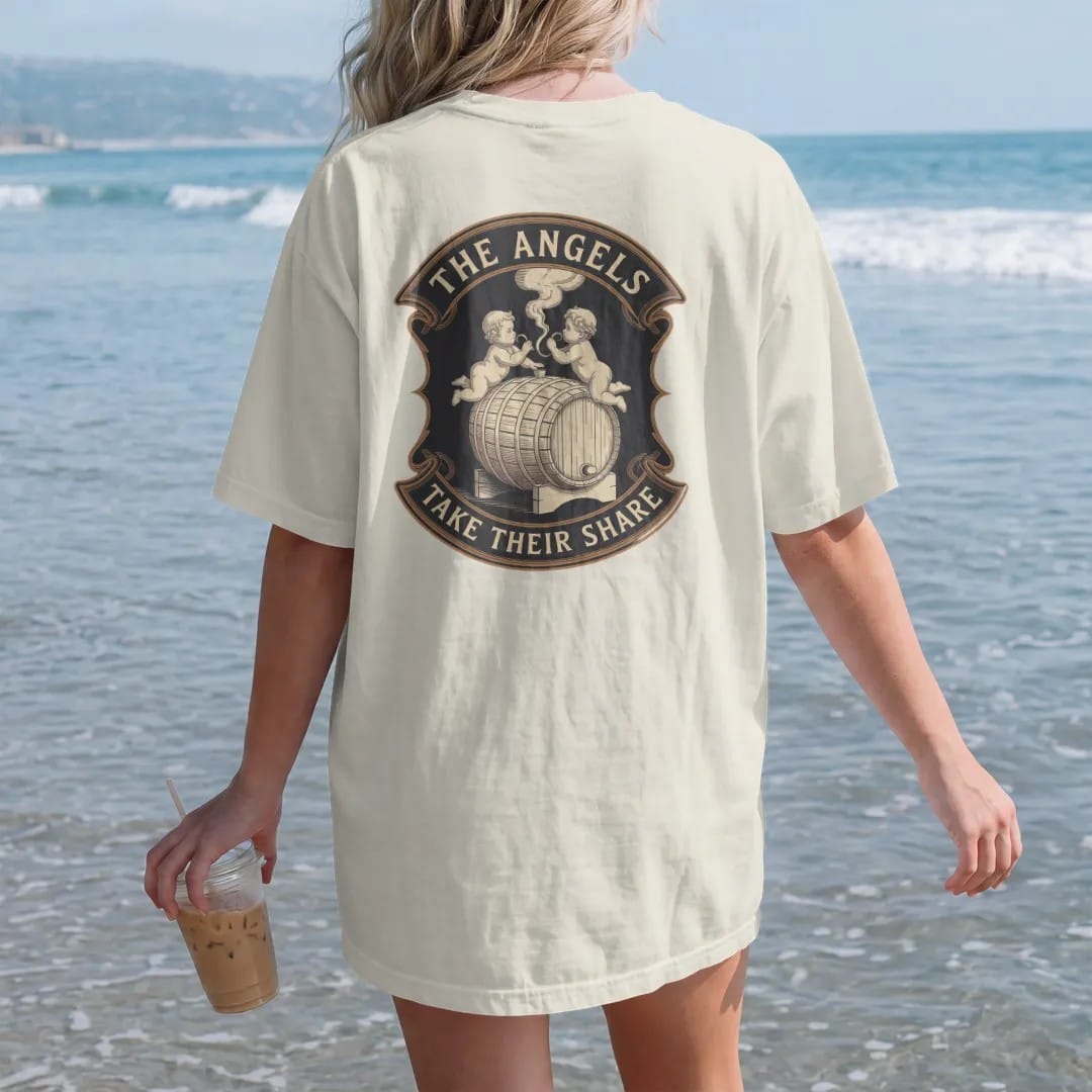 Woman on the beach in a Ivory tee, back view: two engraved cherubs sipping vapor from an oak barrel, with "THE ANGELS" and "TAKE THEIR SHARE" in ornate engraved Didone capitals, printed as a fine engraving in antique gold and warm cream.
