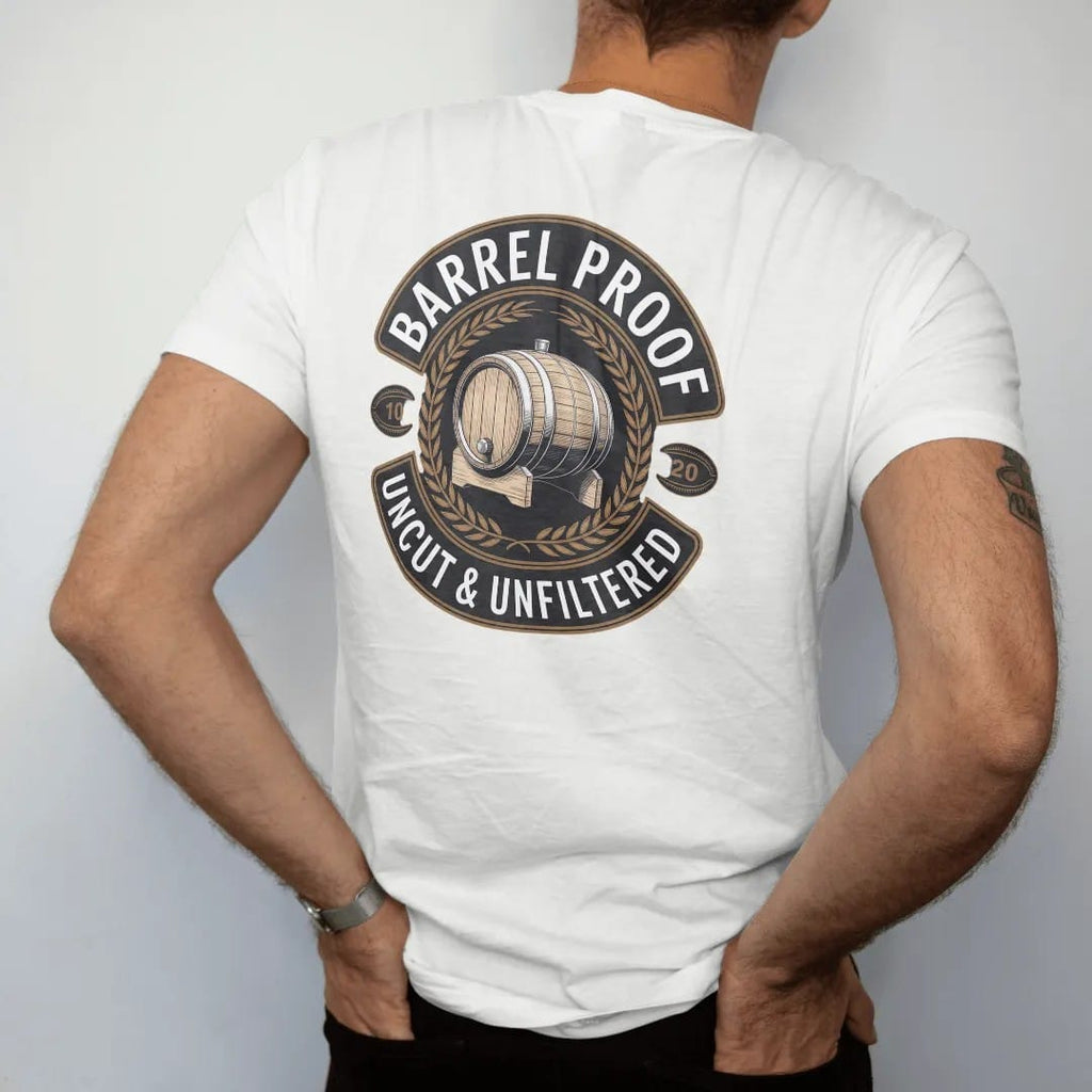 Man wearing a White tee, seen from the back: a round distillery badge with an engraved oak barrel and wheat sprigs, with "BARREL PROOF" and "UNCUT & UNFILTERED" in condensed Victorian display capitals, printed as a fine engraving in antique gold…