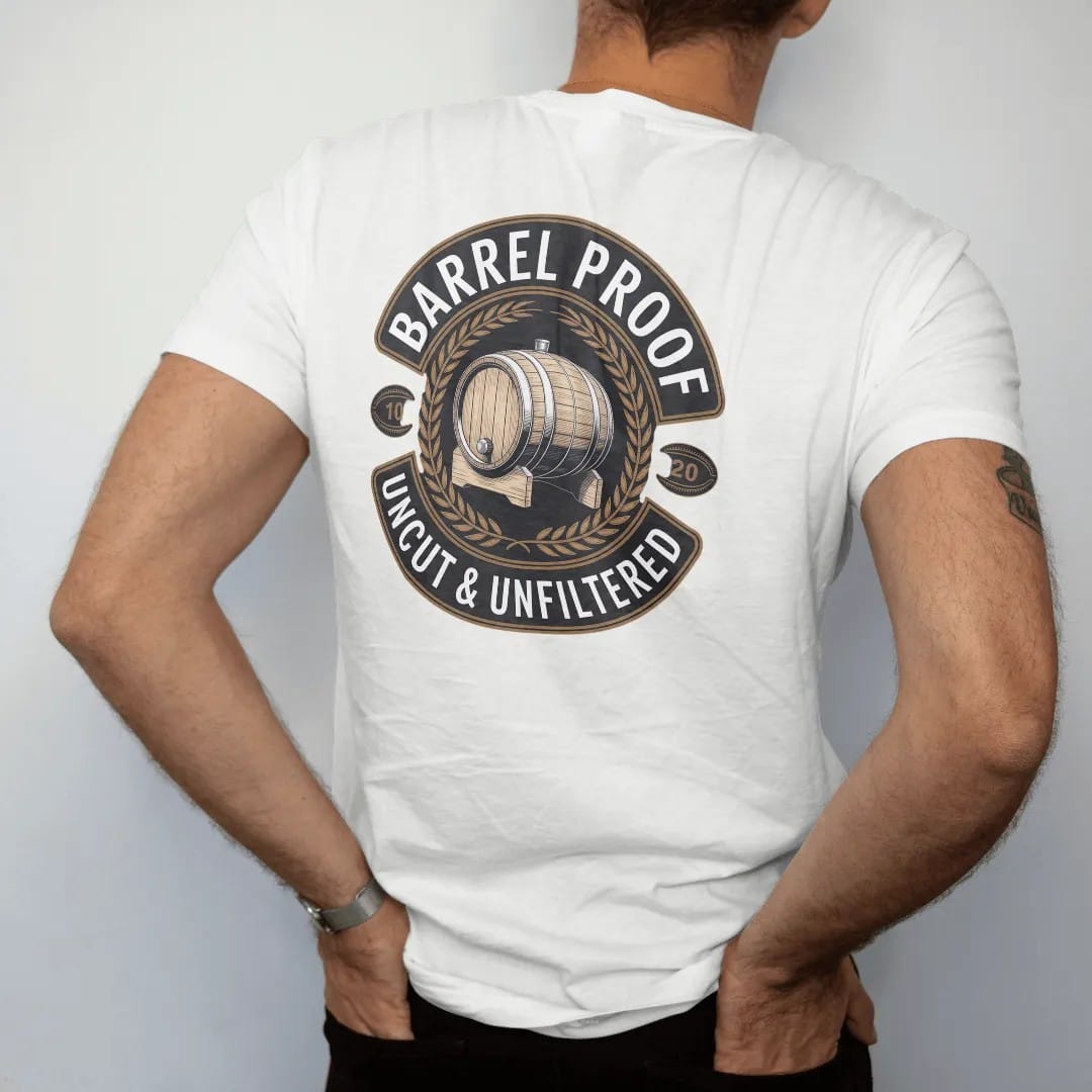 Man wearing a White tee, seen from the back: a round distillery badge with an engraved oak barrel and wheat sprigs, with "BARREL PROOF" and "UNCUT & UNFILTERED" in condensed Victorian display capitals, printed as a fine engraving in antique gold…