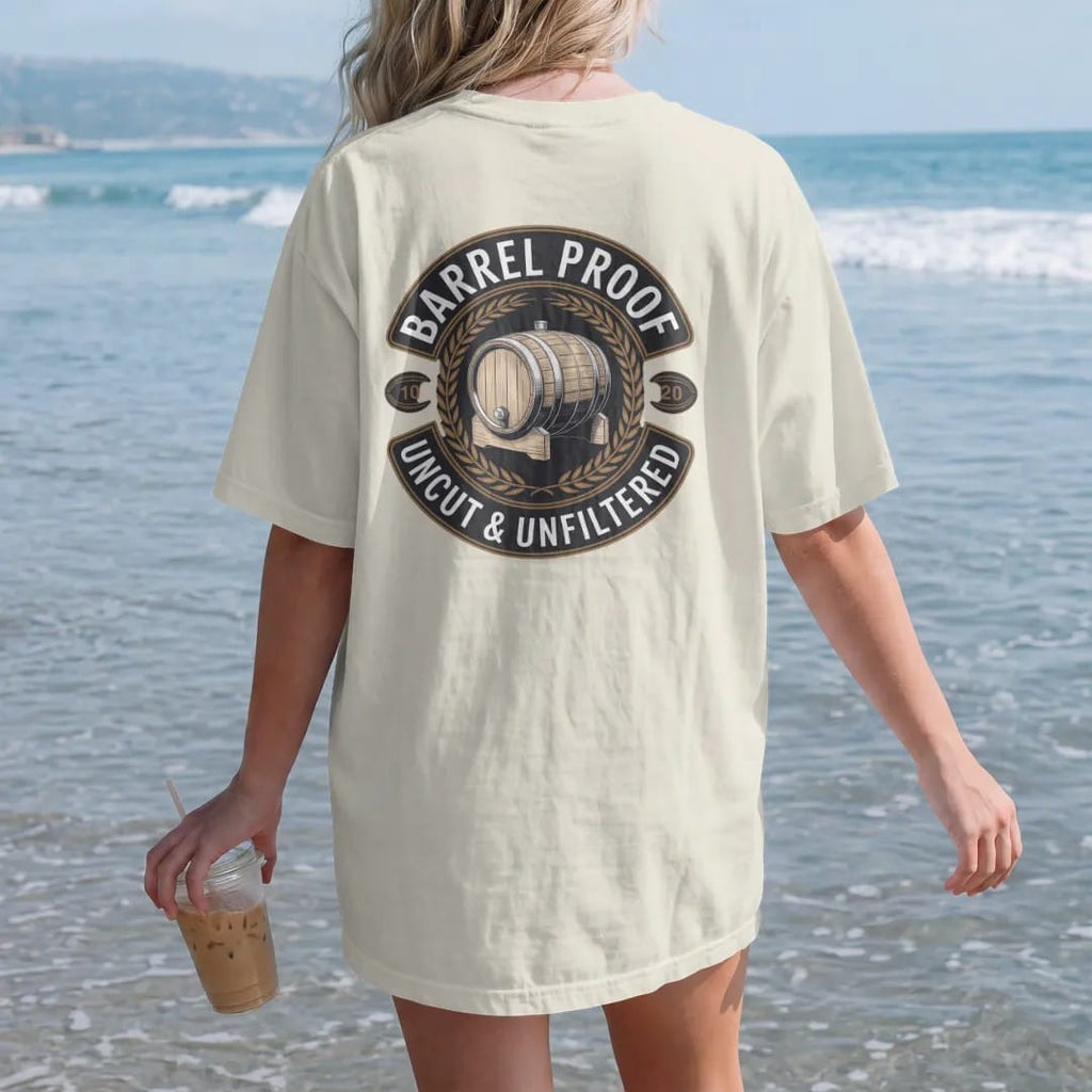 Woman on the beach in a Ivory tee, back view: a round distillery badge with an engraved oak barrel and wheat sprigs, with "BARREL PROOF" and "UNCUT & UNFILTERED" in condensed Victorian display capitals, printed as a fine engraving in antique gold…