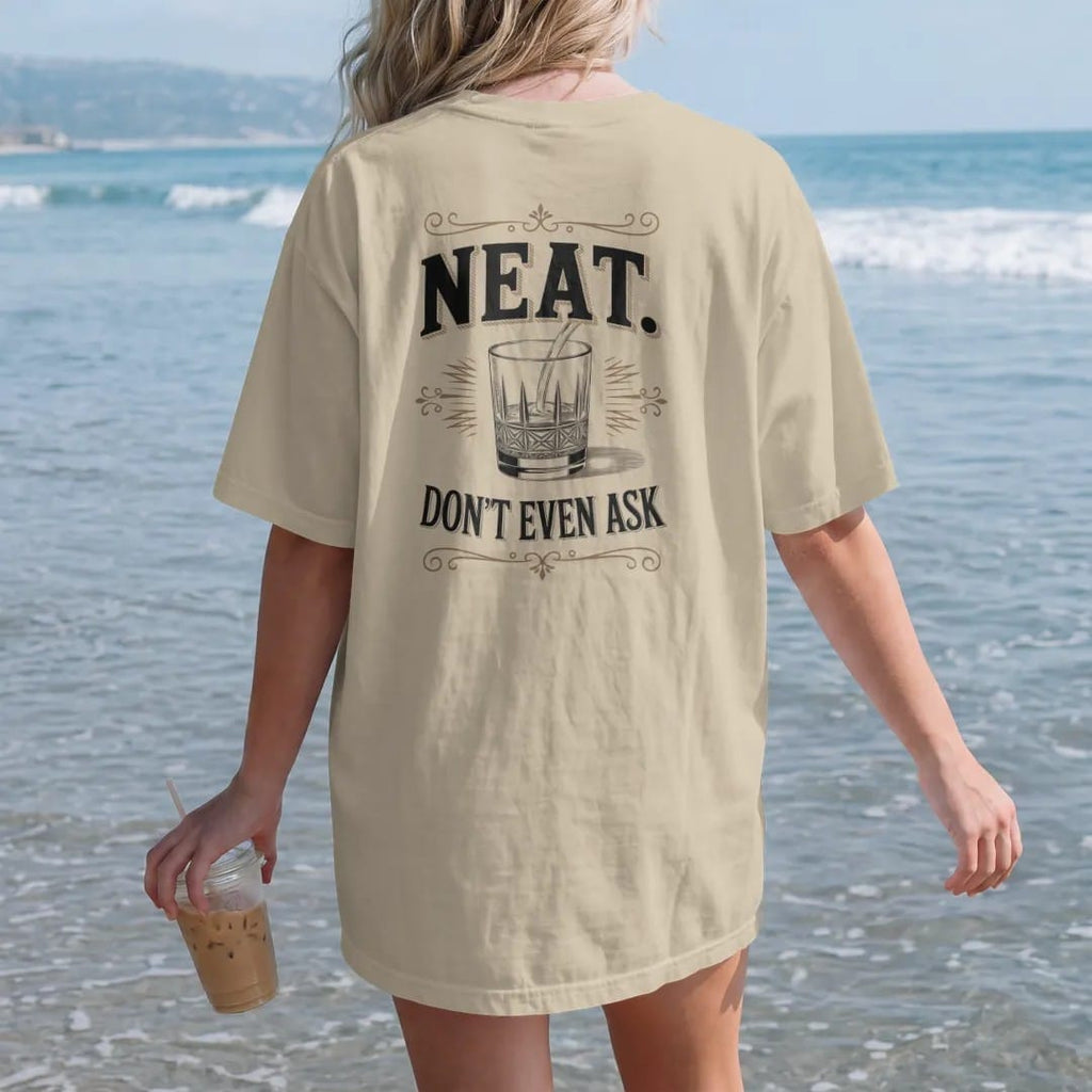 Woman on the beach in a Sandstone tee, back view: an oversized serif word over a small crystal rocks glass, with "NEAT." and "DON'T EVEN ASK" in heavy high-contrast Didone serif with small engraved caps, printed as a fine engraving in oxblood,…