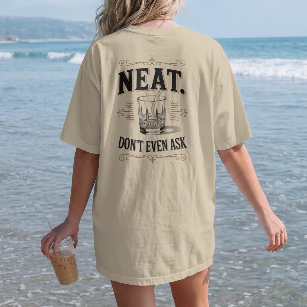 Woman on the beach in a Sandstone tee, back view: an oversized serif word over a small crystal rocks glass, with "NEAT." and "DON'T EVEN ASK" in heavy high-contrast Didone serif with small engraved caps, printed as a fine engraving in oxblood,…