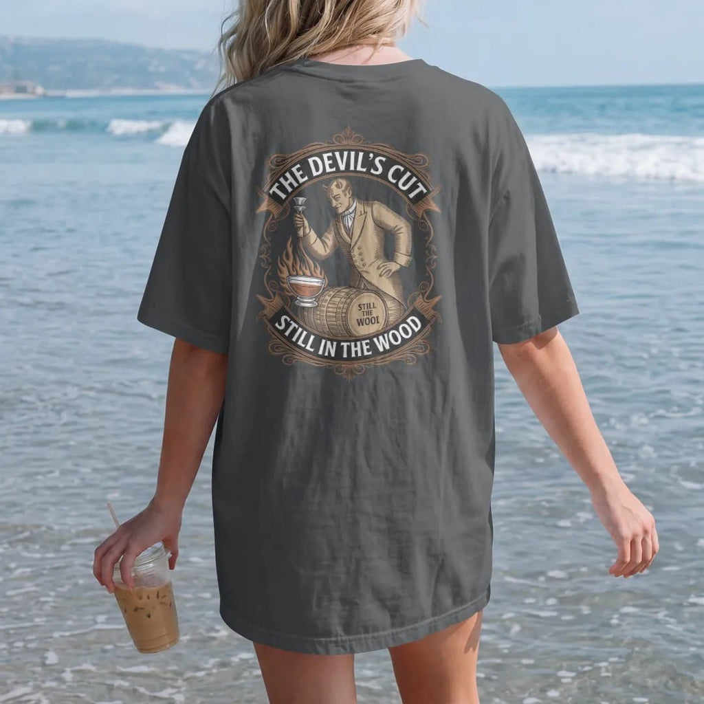 Woman on the beach in a Graphite tee, back view: an engraved devil in a waistcoat wringing whiskey from a barrel stave, with "THE DEVIL'S CUT" and "STILL IN THE WOOD" in ornate Victorian display capitals, printed as a fine engraving in antique gold…