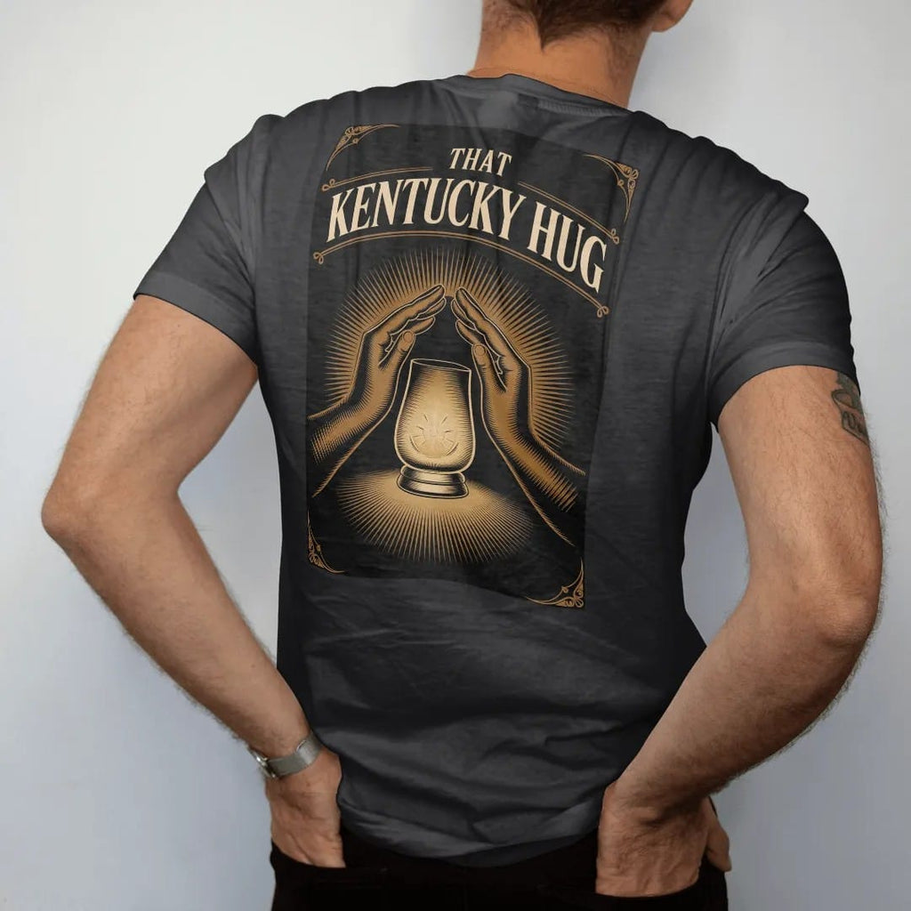 Man wearing a Black tee, seen from the back: a glowing glencairn glass wrapped in two engraved arms, with "THAT KENTUCKY HUG" in warm rounded Victorian serif capitals, printed as a fine engraving in antique gold and warm cream.