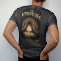 Man wearing a Black tee, seen from the back: a glowing glencairn glass wrapped in two engraved arms, with "THAT KENTUCKY HUG" in warm rounded Victorian serif capitals, printed as a fine engraving in antique gold and warm cream.