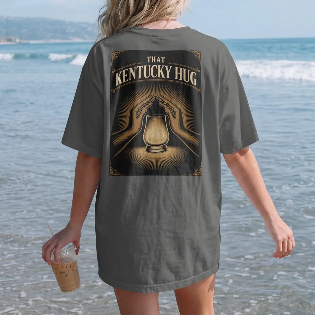 Woman on the beach in a Pepper tee, back view: a glowing glencairn glass wrapped in two engraved arms, with "THAT KENTUCKY HUG" in warm rounded Victorian serif capitals, printed as a fine engraving in antique gold and warm cream.