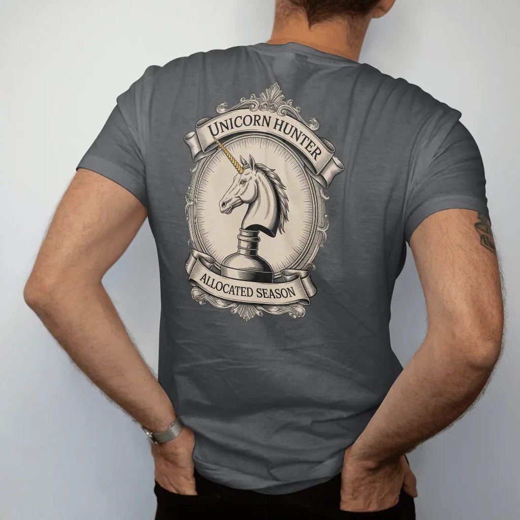Man wearing a Graphite tee, seen from the back: an engraved unicorn head emerging from a whiskey bottle, with "UNICORN HUNTER" and "ALLOCATED SEASON" in ornate engraved Didone capitals, printed as a fine engraving in antique gold and warm cream.