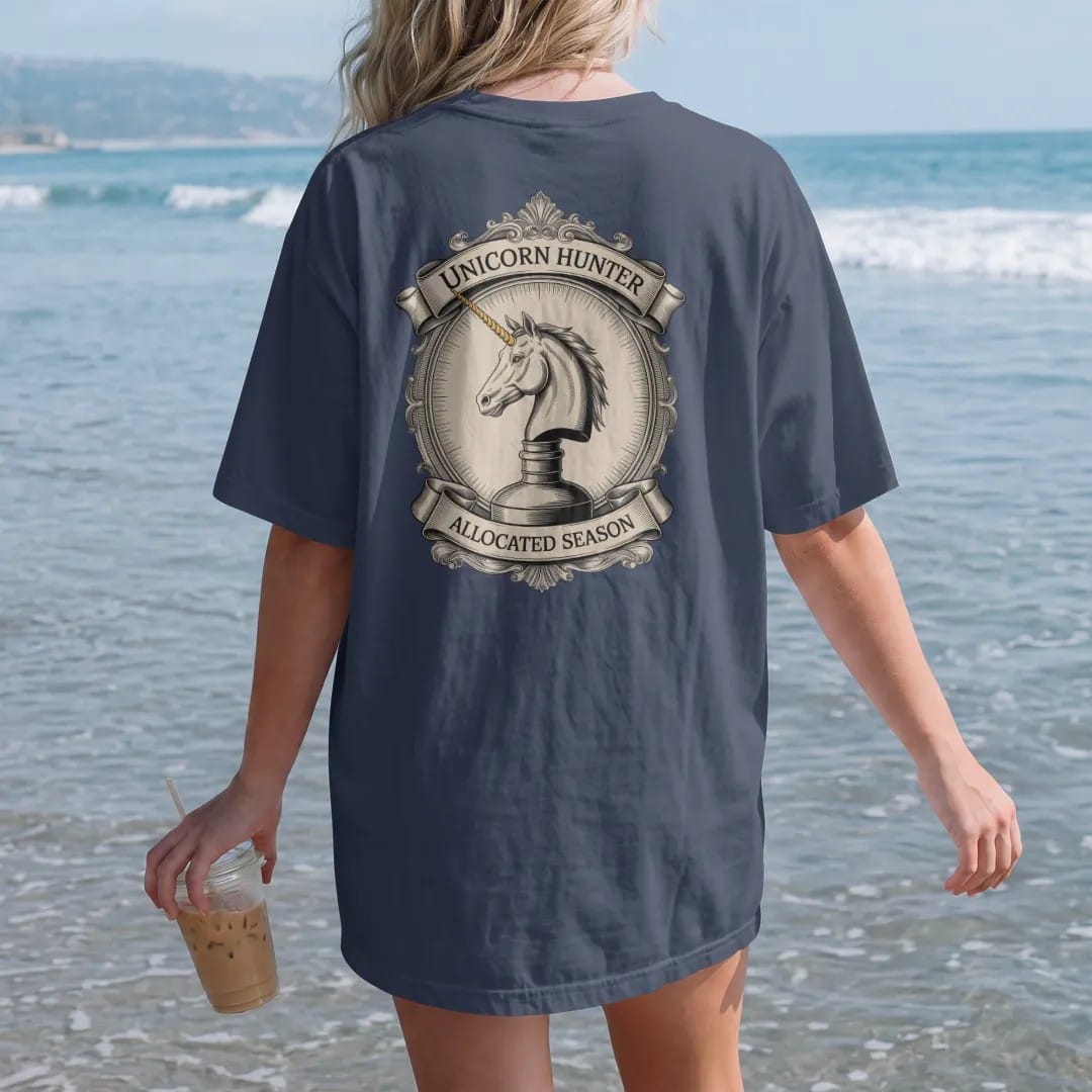 Woman on the beach in a Navy tee, back view: an engraved unicorn head emerging from a whiskey bottle, with "UNICORN HUNTER" and "ALLOCATED SEASON" in ornate engraved Didone capitals, printed as a fine engraving in antique gold and warm cream.