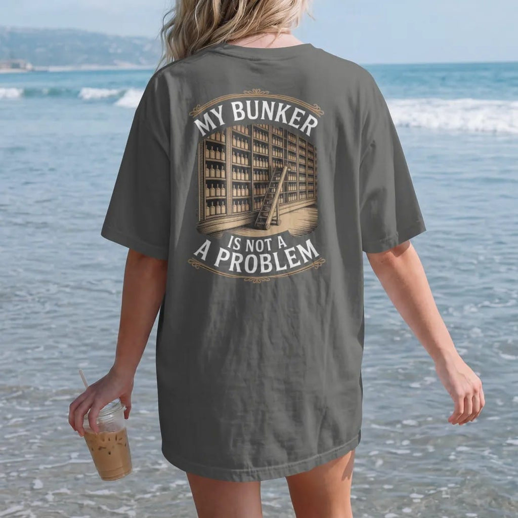 Woman on the beach in a Pepper tee, back view: a wall of shelves packed with whiskey bottles and a library ladder, with "MY BUNKER" and "IS NOT A PROBLEM", printed as a fine engraving in antique gold and warm cream.