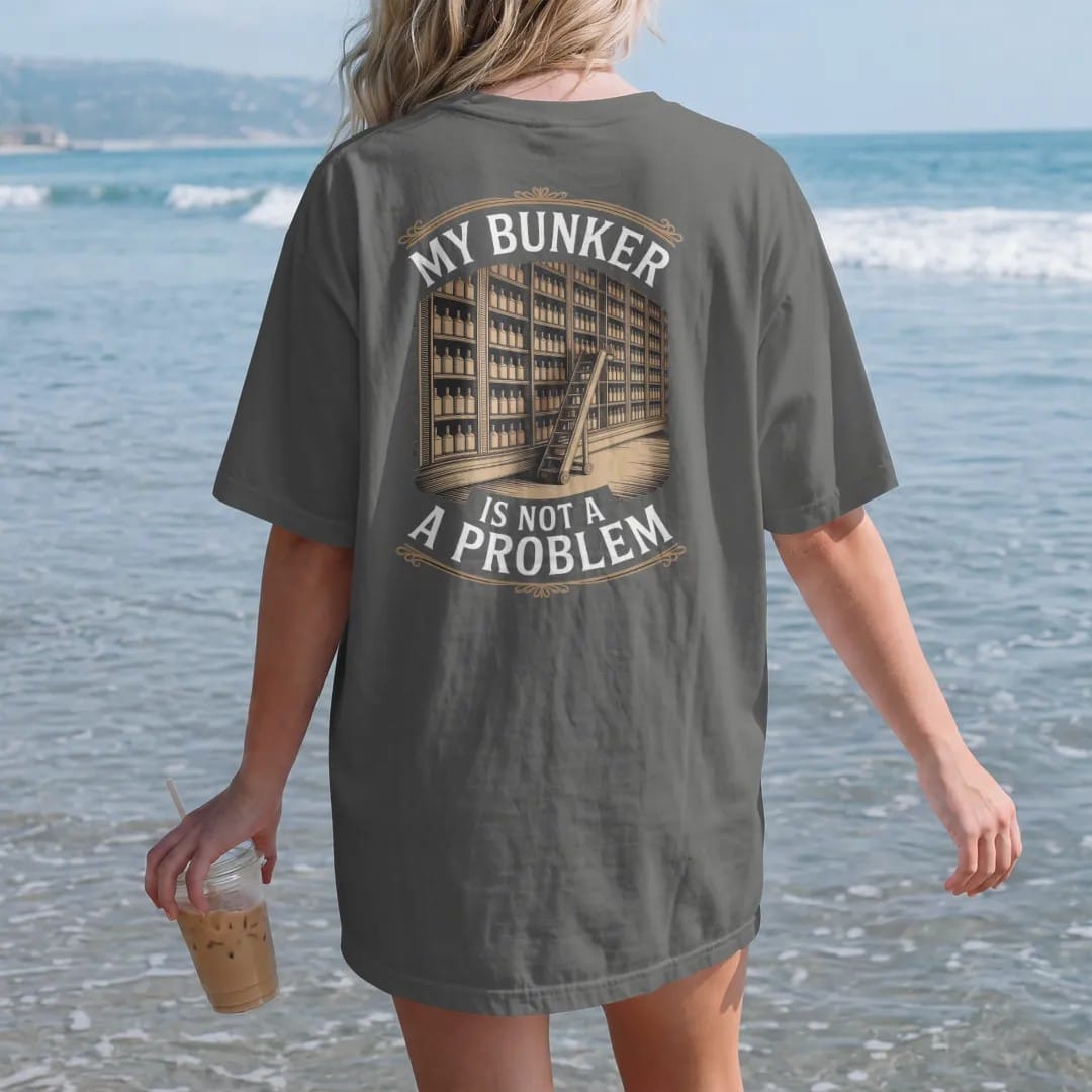 Woman on the beach in a Pepper tee, back view: a wall of shelves packed with whiskey bottles and a library ladder, with "MY BUNKER" and "IS NOT A PROBLEM", printed as a fine engraving in antique gold and warm cream.