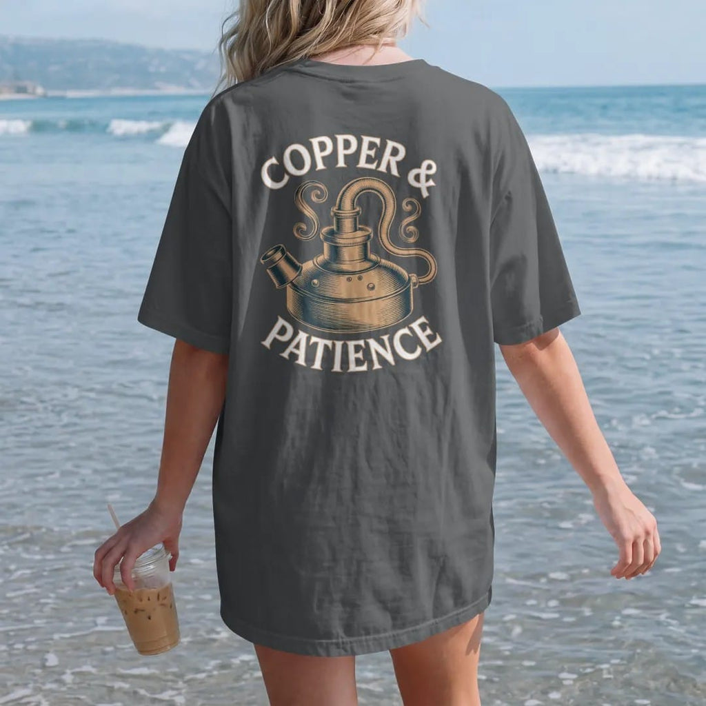 Woman on the beach in a Graphite tee, back view: an engraved copper pot still with swan neck and condenser, with "COPPER & PATIENCE", printed as a fine engraving in antique gold and warm cream.