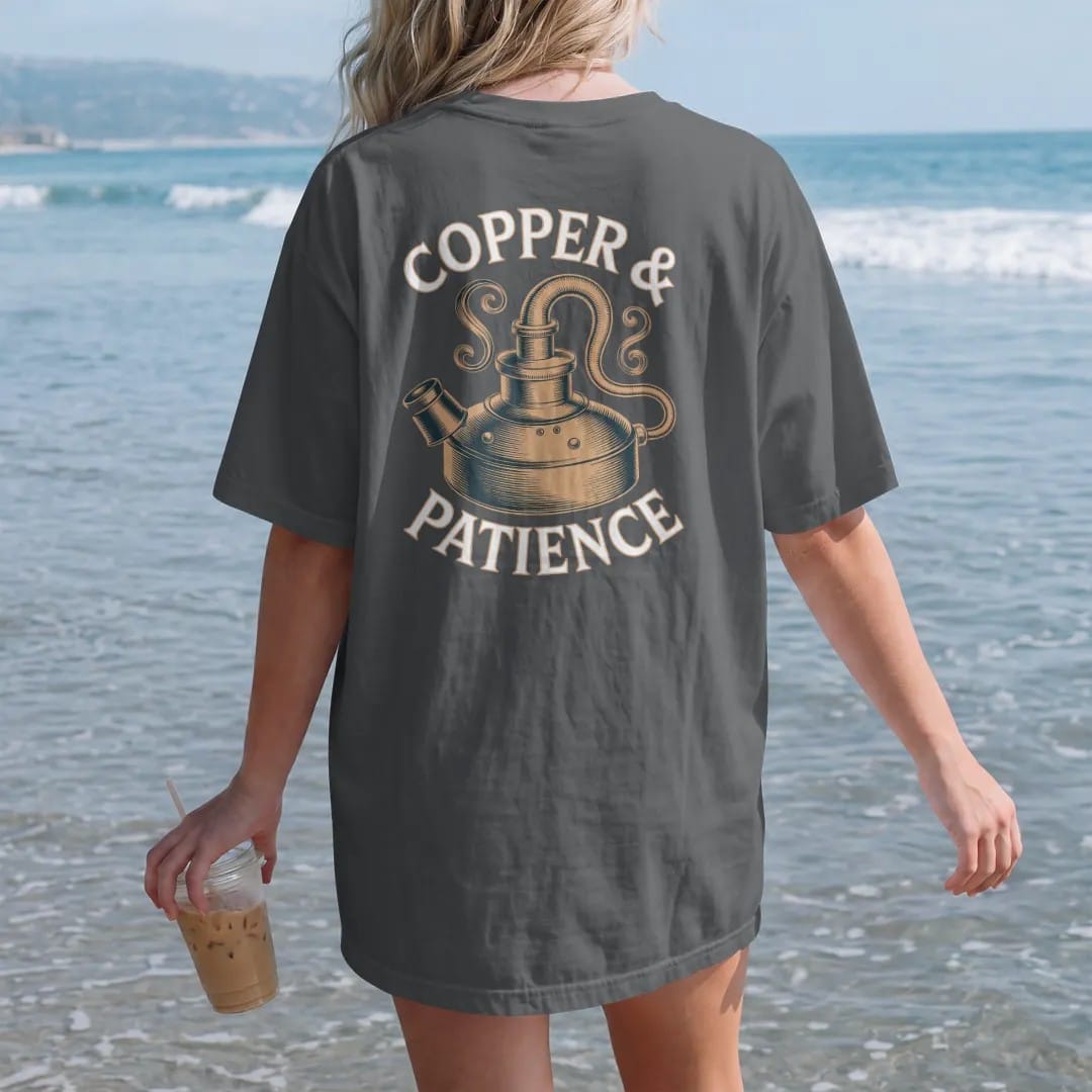 Woman on the beach in a Graphite tee, back view: an engraved copper pot still with swan neck and condenser, with "COPPER & PATIENCE", printed as a fine engraving in antique gold and warm cream.