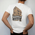 Man wearing a White tee, seen from the back: an engraved wooden rickhouse barn full of barrels, with "RICKHOUSE" and "TOP FLOOR AGED" in condensed Victorian display capitals, printed as a fine engraving in antique gold and warm cream.