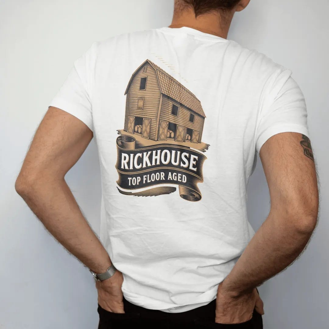Man wearing a White tee, seen from the back: an engraved wooden rickhouse barn full of barrels, with "RICKHOUSE" and "TOP FLOOR AGED" in condensed Victorian display capitals, printed as a fine engraving in antique gold and warm cream.