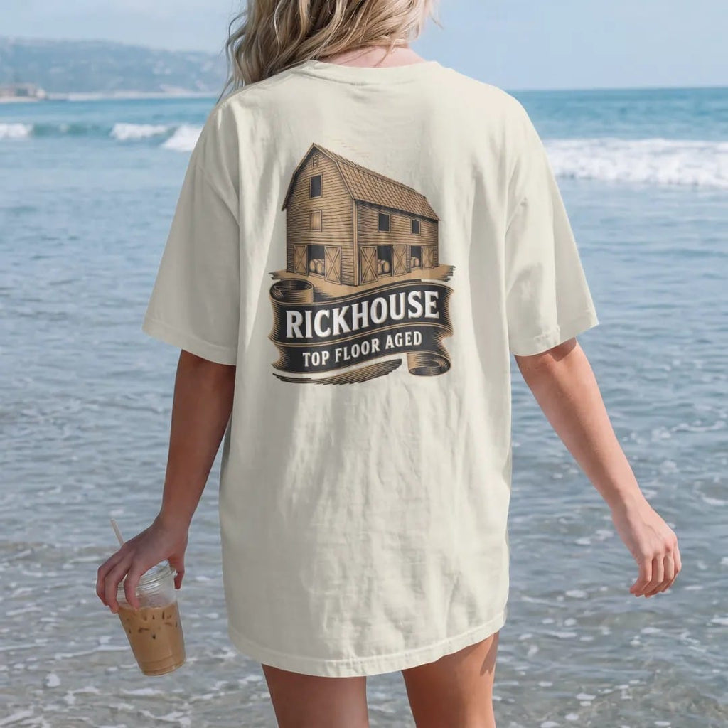 Woman on the beach in a Ivory tee, back view: an engraved wooden rickhouse barn full of barrels, with "RICKHOUSE" and "TOP FLOOR AGED" in condensed Victorian display capitals, printed as a fine engraving in antique gold and warm cream.