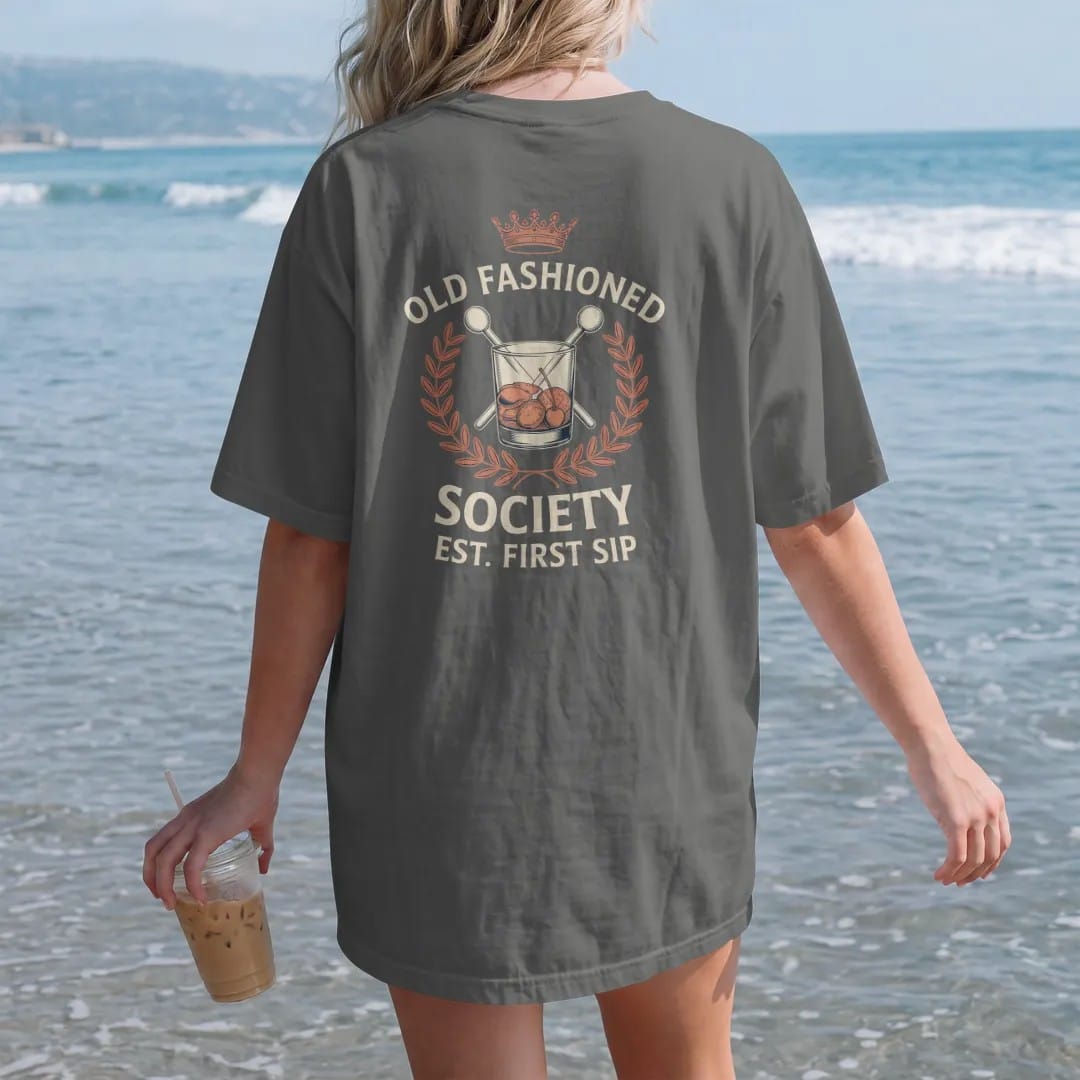 Woman on the beach in a Pepper tee, back view: a members-club crest with an Old Fashioned glass and crossed muddlers, with "OLD FASHIONED", "SOCIETY" and "EST. FIRST SIP" in engraved serif small caps with a Didone headline, printed as a fine…
