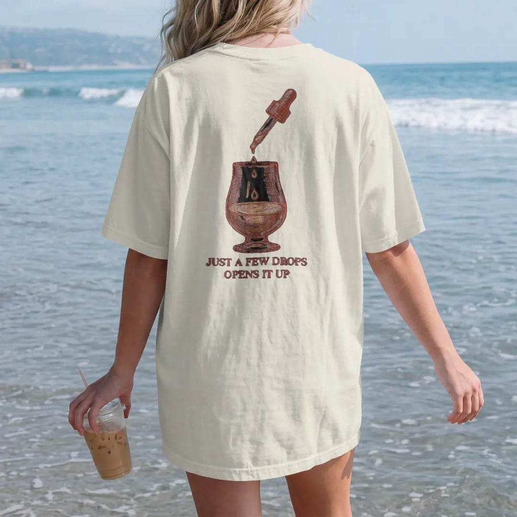 Woman on the beach in a Ivory tee, back view: a glass dropper releasing drops into a glencairn glass, with "JUST A FEW DROPS" and "OPENS IT UP", printed as a fine engraving in oxblood, charred oak brown and antique gold.