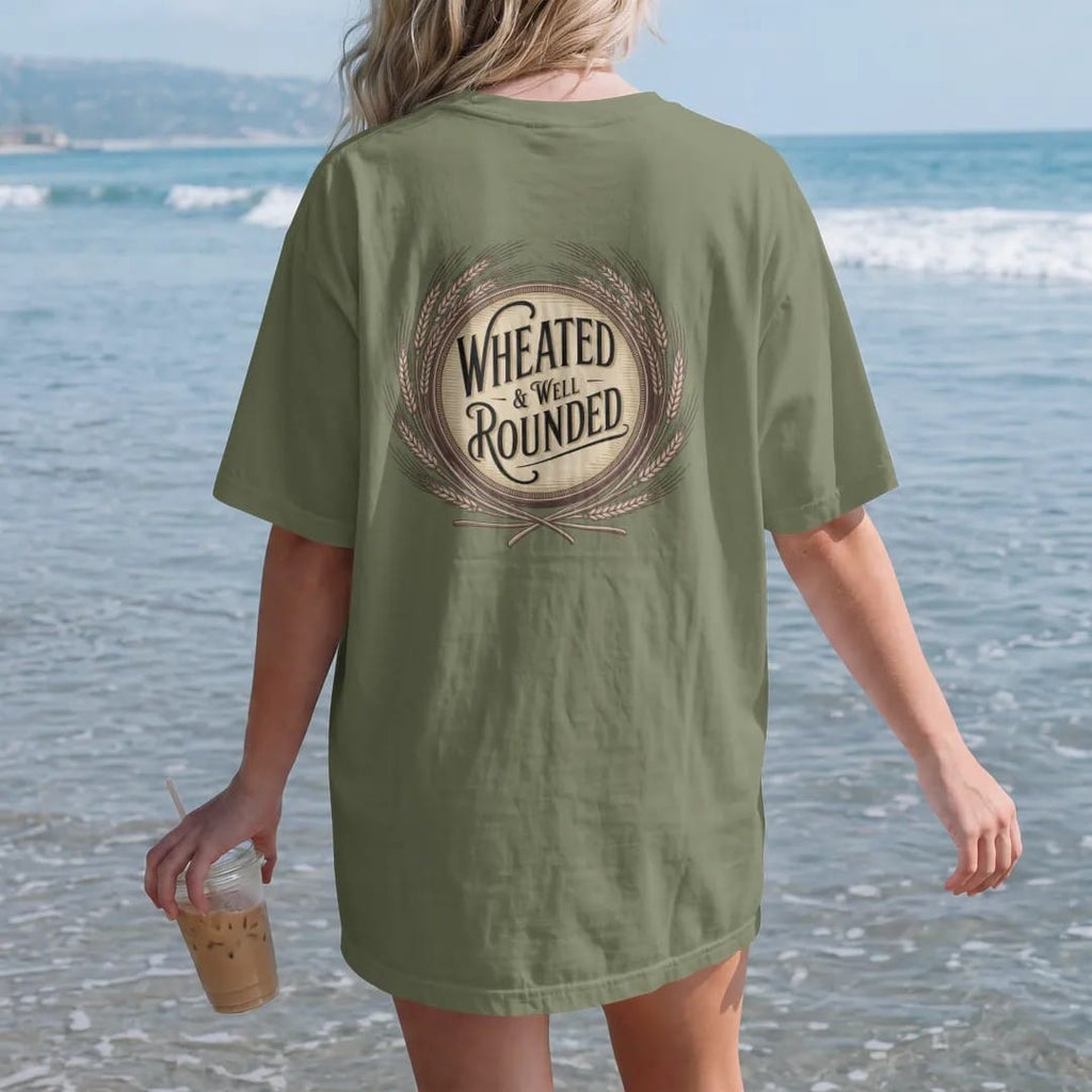 Woman on the beach in a Moss tee, back view: a wreath of wheat stalks around a round barrel head, with "WHEATED" and "& WELL ROUNDED" in elegant Didone capitals with copperplate script, printed as a fine engraving in antique gold and warm cream.
