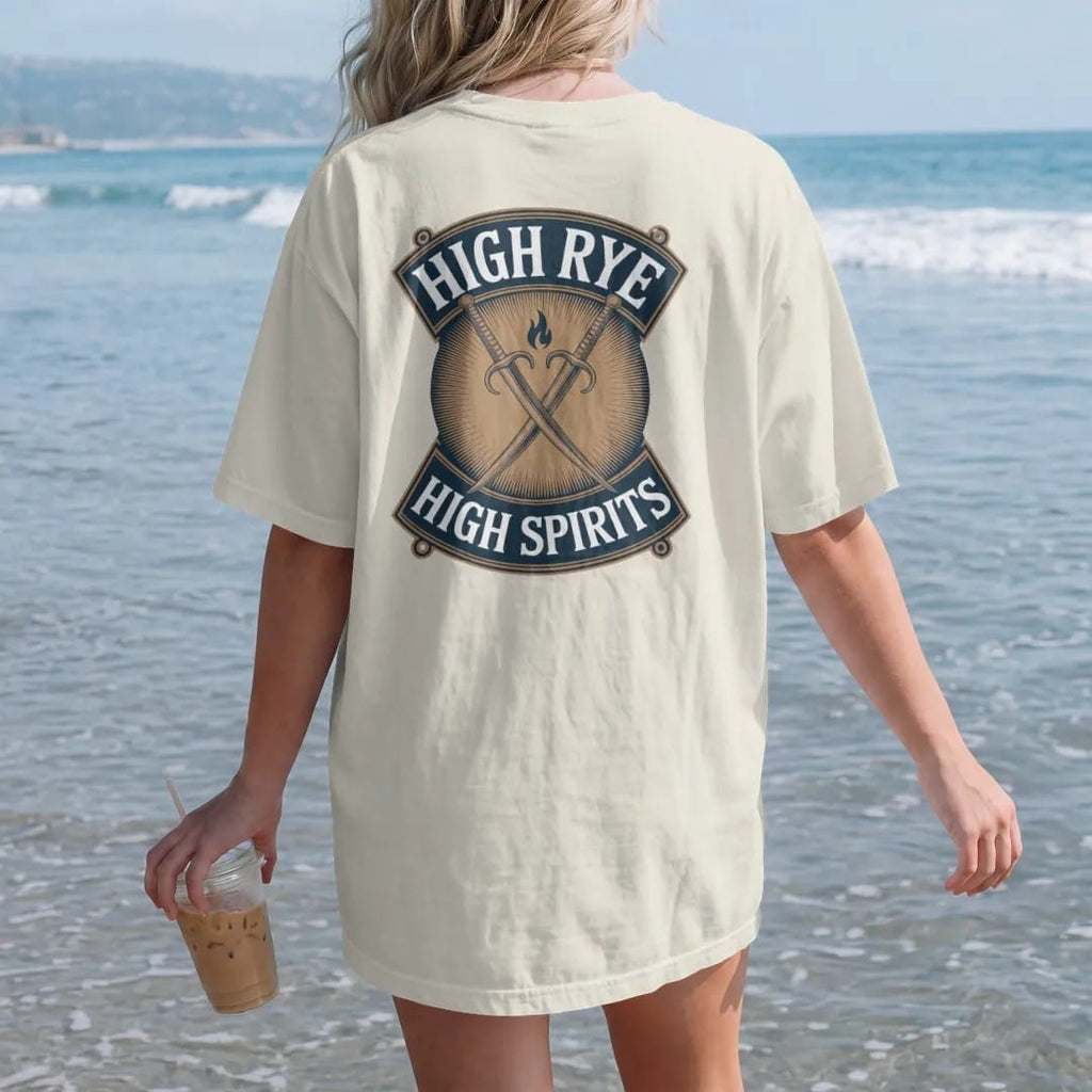 Woman on the beach in a Ivory tee, back view: crossed rye stalks behind a small flame on a shield, with "HIGH RYE" and "HIGH SPIRITS" in condensed Victorian display capitals, printed as a fine engraving in antique gold and warm cream.