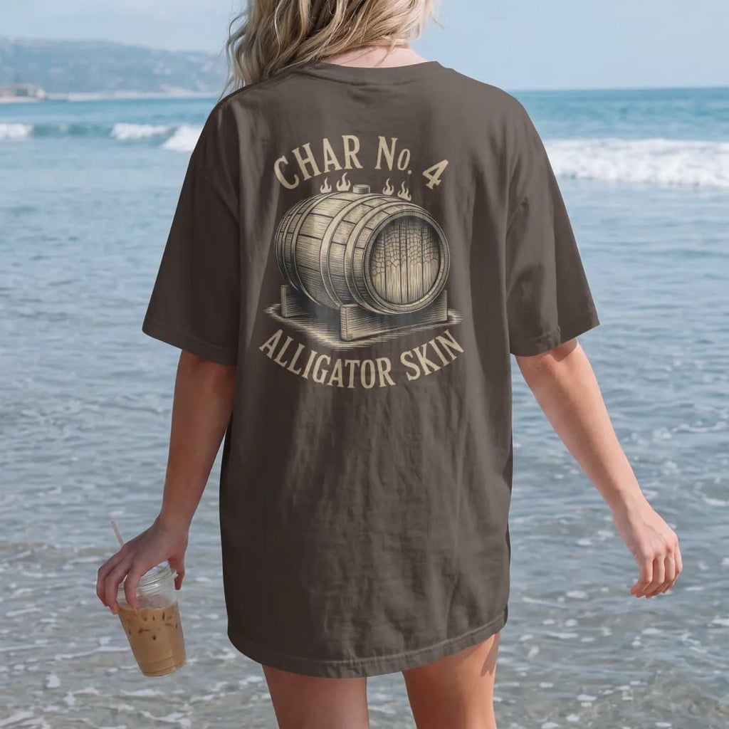 Woman on the beach in a Espresso tee, back view: a cutaway barrel interior with cracked alligator-skin char, with "CHAR NO. 4" and "ALLIGATOR SKIN", printed as a fine engraving in antique gold and warm cream.