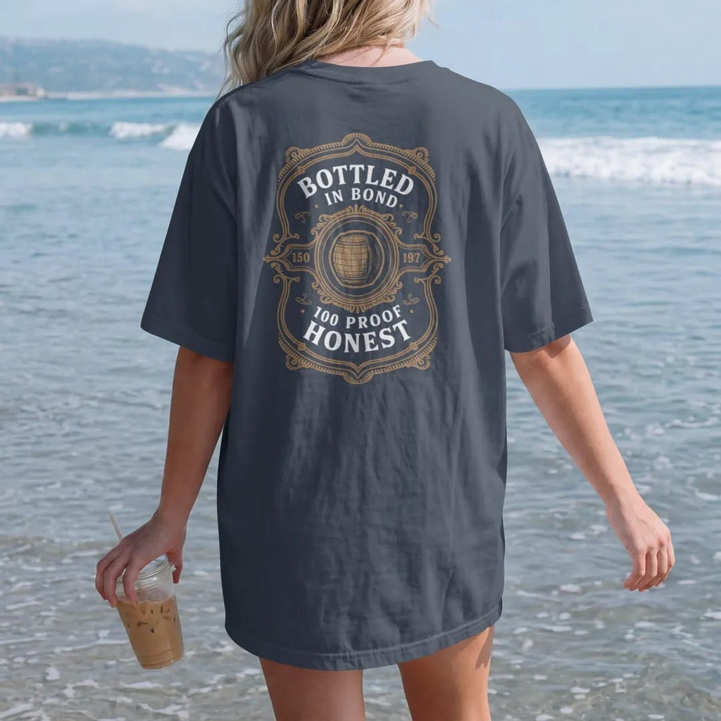 Woman on the beach in a Midnight tee, back view: a guilloche-engraved tax-strip emblem with a barrel icon, with "BOTTLED IN BOND" and "100 PROOF HONEST", printed as a fine engraving in antique gold and warm cream.