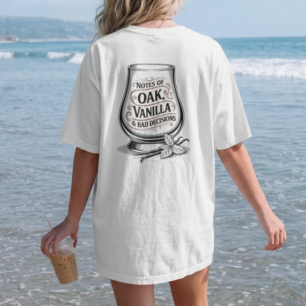 Woman on the beach in a White tee, back view: a glencairn glass with swirling aroma wisps, with "NOTES OF", "OAK, VANILLA" and "& BAD DECISIONS", printed as a fine engraving in oxblood, charred oak brown and antique gold.