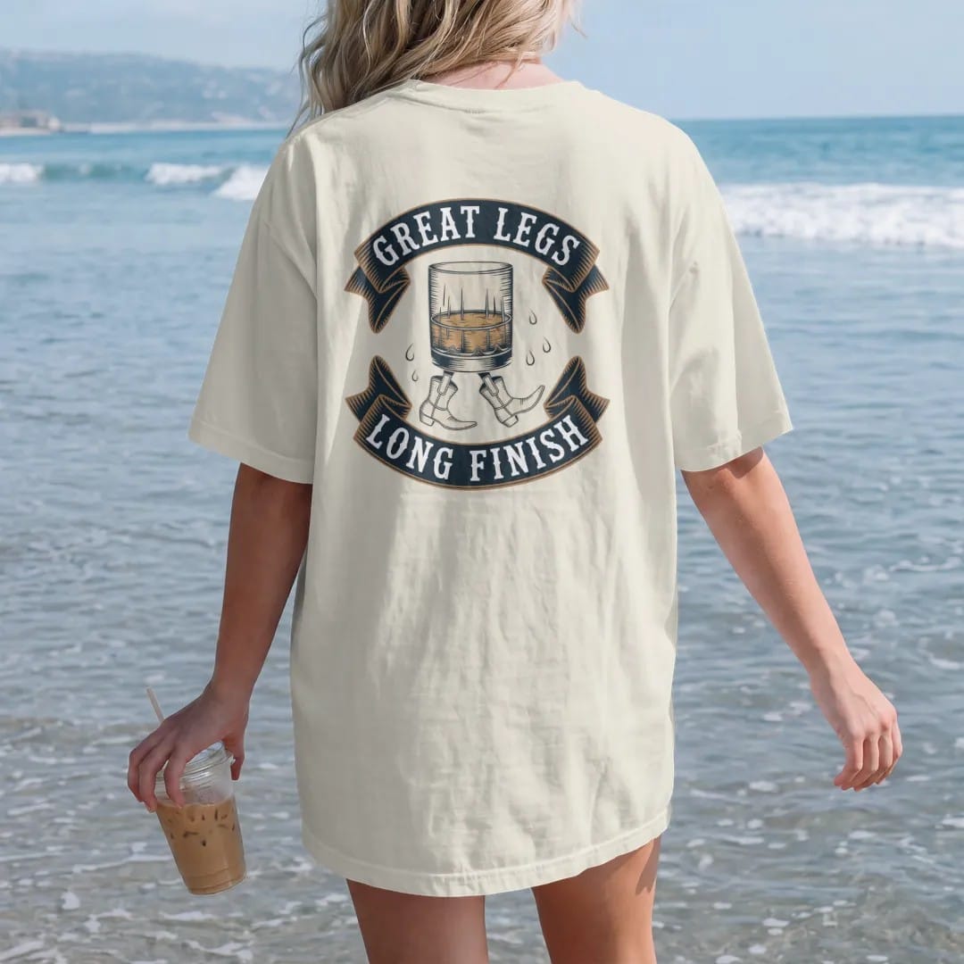 Woman on the beach in a Ivory tee, back view: a whiskey glass walking on cartoon legs in cowboy boots, with "GREAT LEGS" and "LONG FINISH" in playful Victorian display capitals, printed as a fine engraving in antique gold and warm cream.