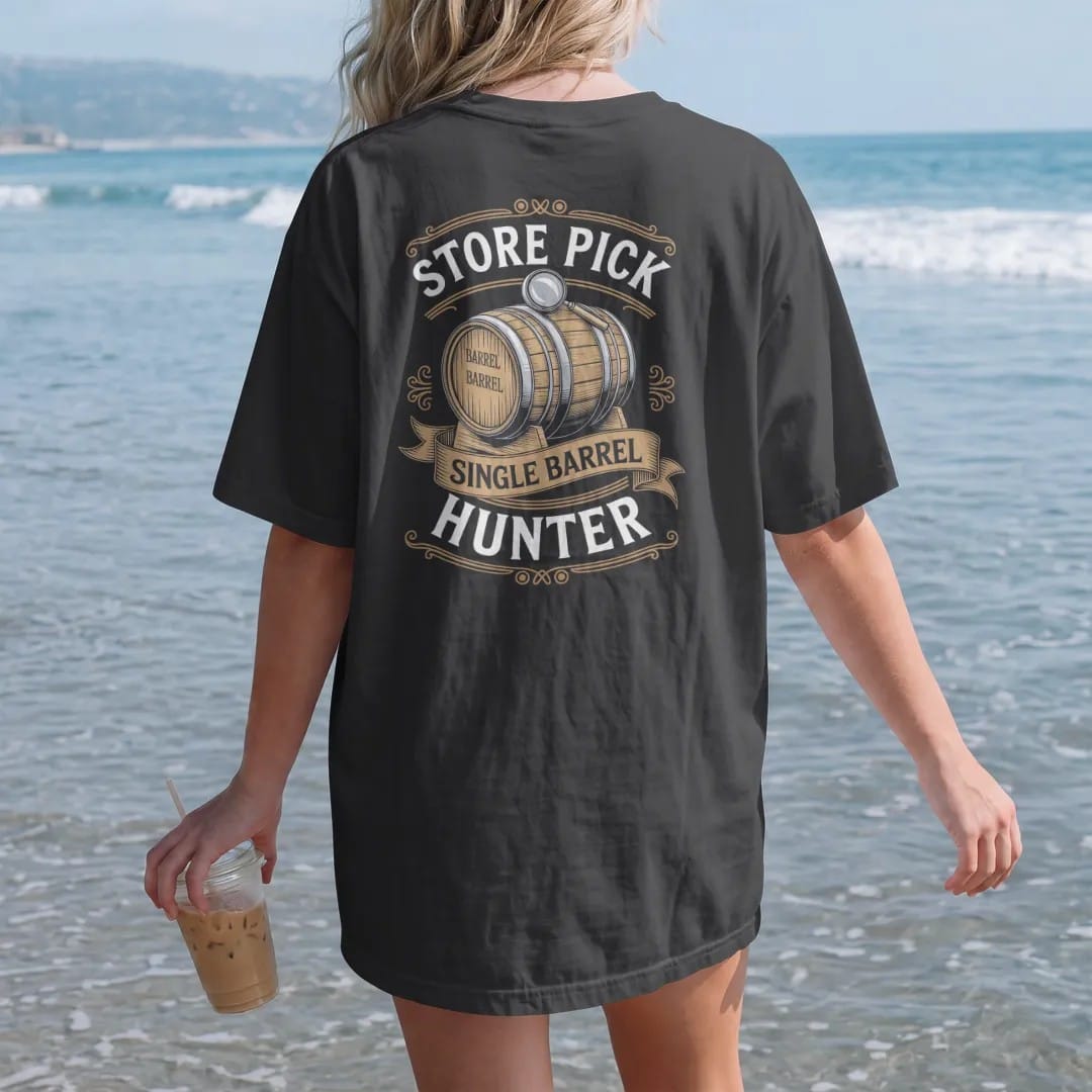 Woman on the beach in a Black tee, back view: an engraved barrel with a chalk number and a magnifying glass, with "STORE PICK" and "SINGLE BARREL HUNTER", printed as a fine engraving in antique gold and warm cream.