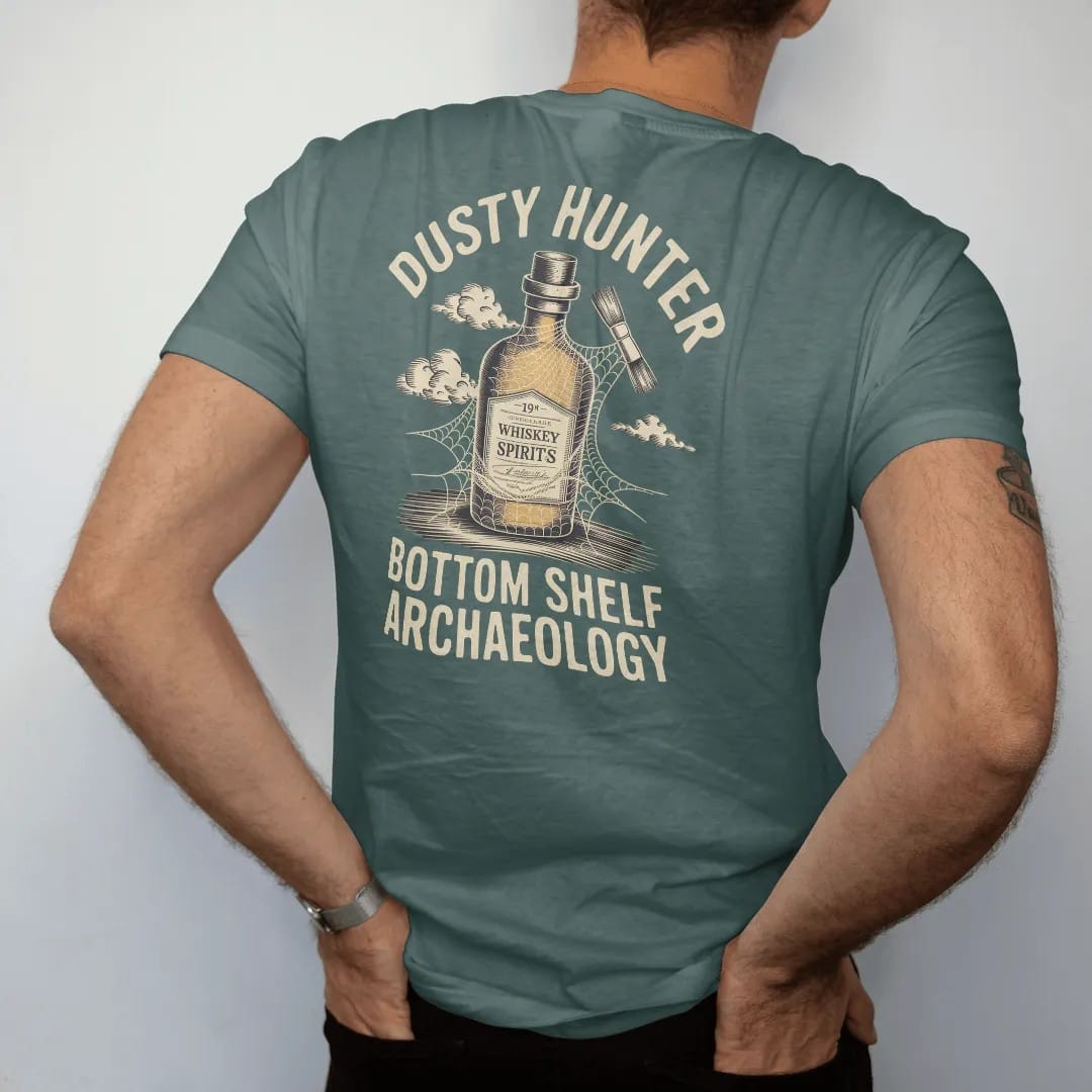 Man wearing a Blue Spruce tee, seen from the back: a cobwebbed whiskey bottle being brushed off with an archaeologist's brush, with "DUSTY HUNTER" and "BOTTOM SHELF ARCHAEOLOGY", printed as a fine engraving in antique gold and warm cream.