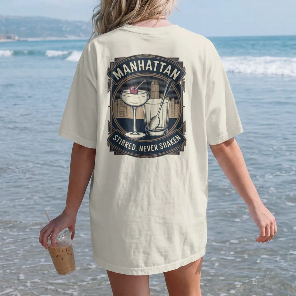 Woman on the beach in a Ivory tee, back view: a coupe with a cherry beside a mixing glass and bar spoon, with "MANHATTAN" and "STIRRED, NEVER SHAKEN" in Art Deco high-contrast serif capitals, printed as a fine engraving in antique gold and warm…