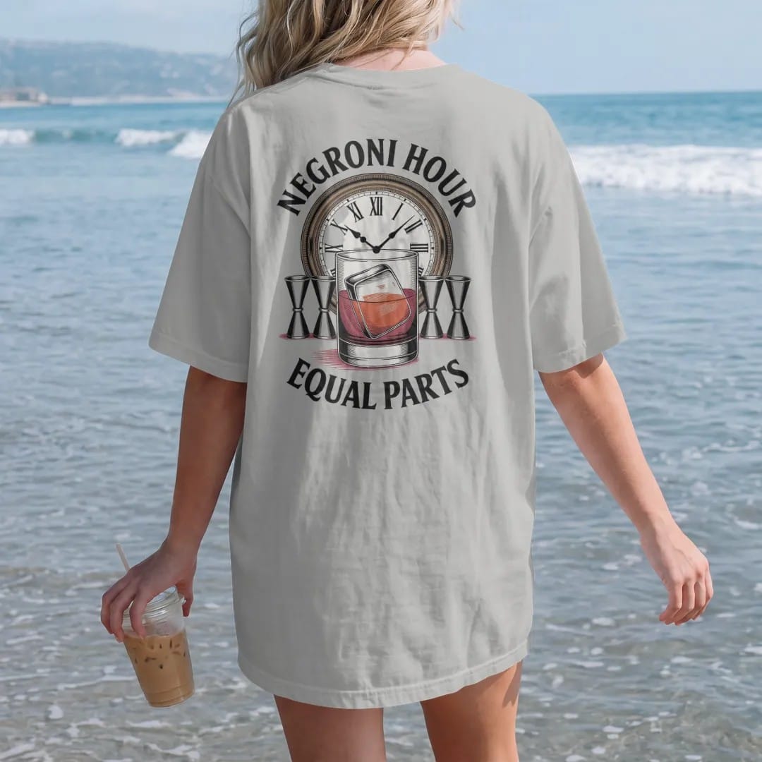 Woman on the beach in a Granite tee, back view: a Negroni glass with orange wheel, three jiggers and a clock face, with "NEGRONI HOUR" and "EQUAL PARTS", printed as a fine engraving in oxblood, charred oak brown and antique gold.