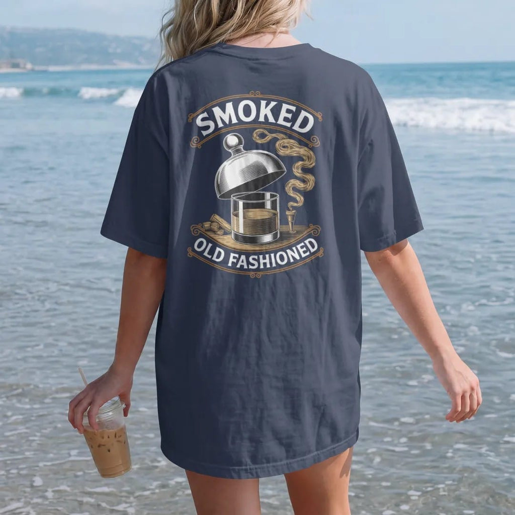 Woman on the beach in a Navy tee, back view: a glass cloche lifting off a rocks glass with curling smoke, with "SMOKED" and "OLD FASHIONED" in elegant Didone capitals, printed as a fine engraving in antique gold and warm cream.