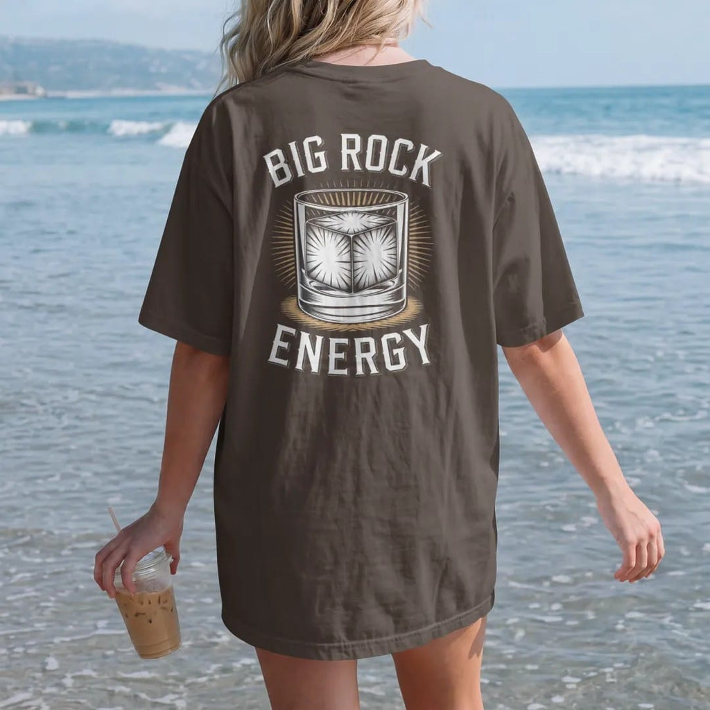 Woman on the beach in a Espresso tee, back view: a huge clear ice cube in a rocks glass, with "BIG ROCK" and "ENERGY" in heavy Didone serif capitals, printed as a fine engraving in antique gold and warm cream.