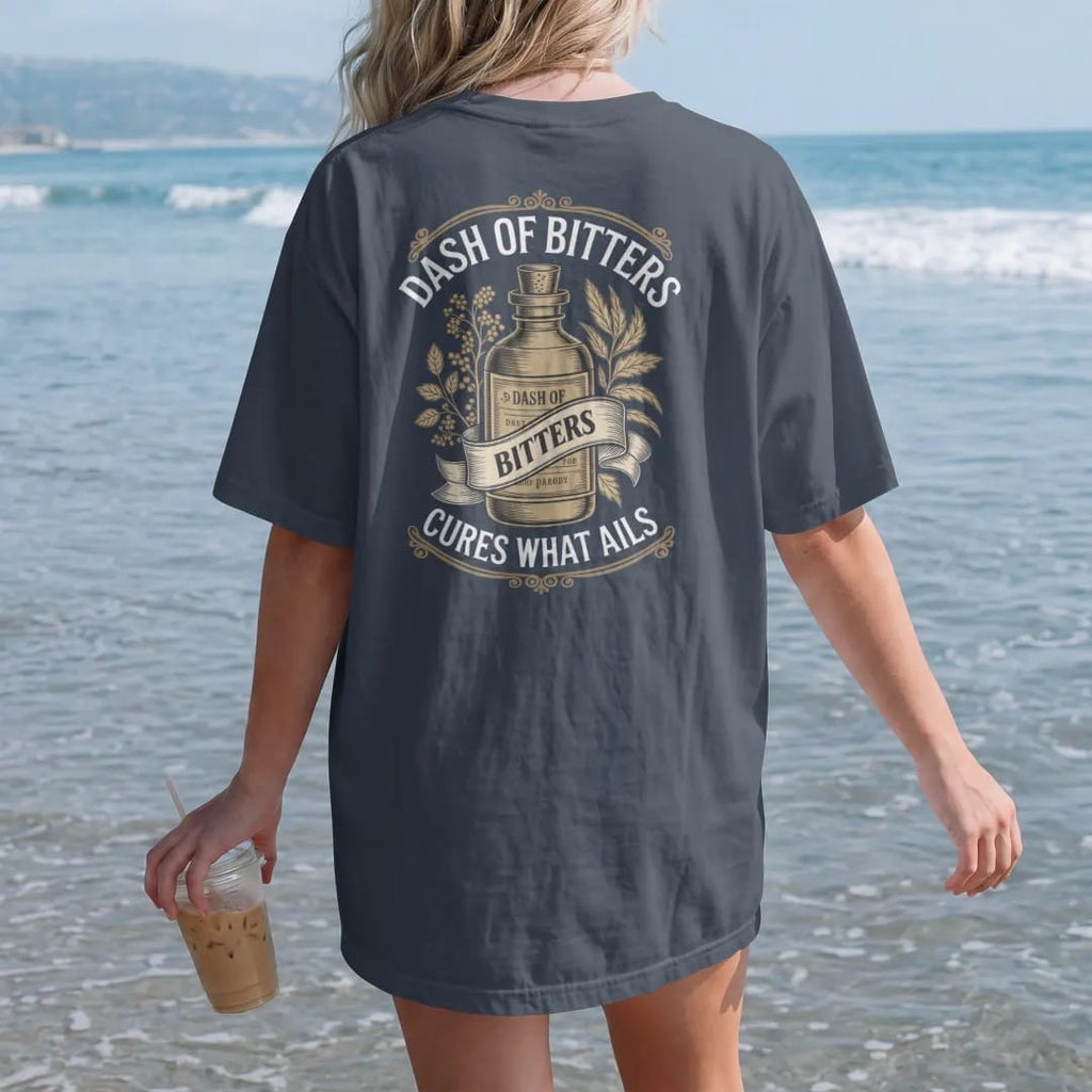 Woman on the beach in a Midnight tee, back view: a paper-wrapped bitters bottle among engraved herbs, with "DASH OF BITTERS" and "CURES WHAT AILS" in Victorian apothecary display serif, printed as a fine engraving in antique gold and warm cream.