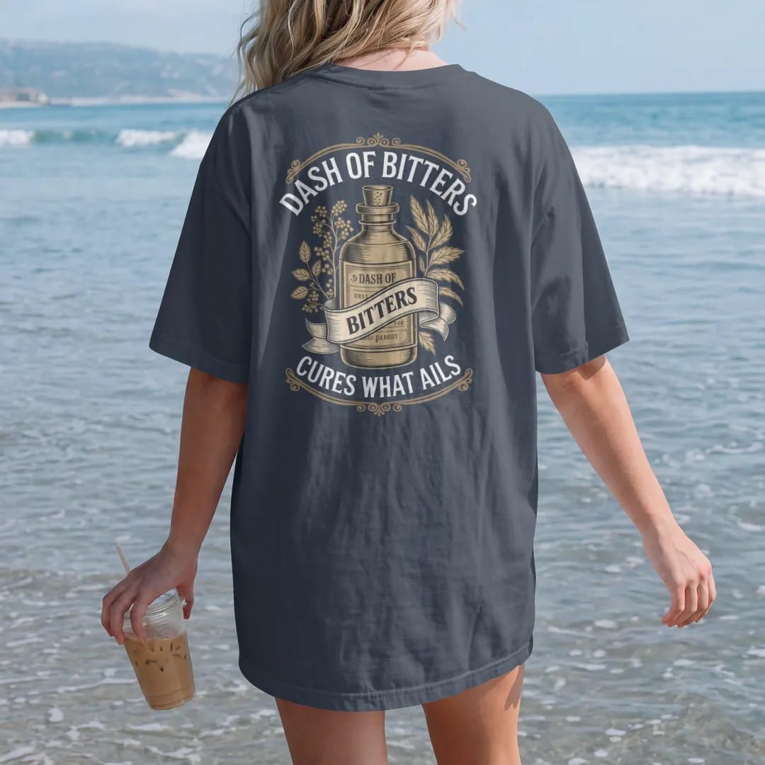 Woman on the beach in a Midnight tee, back view: a paper-wrapped bitters bottle among engraved herbs, with "DASH OF BITTERS" and "CURES WHAT AILS" in Victorian apothecary display serif, printed as a fine engraving in antique gold and warm cream.