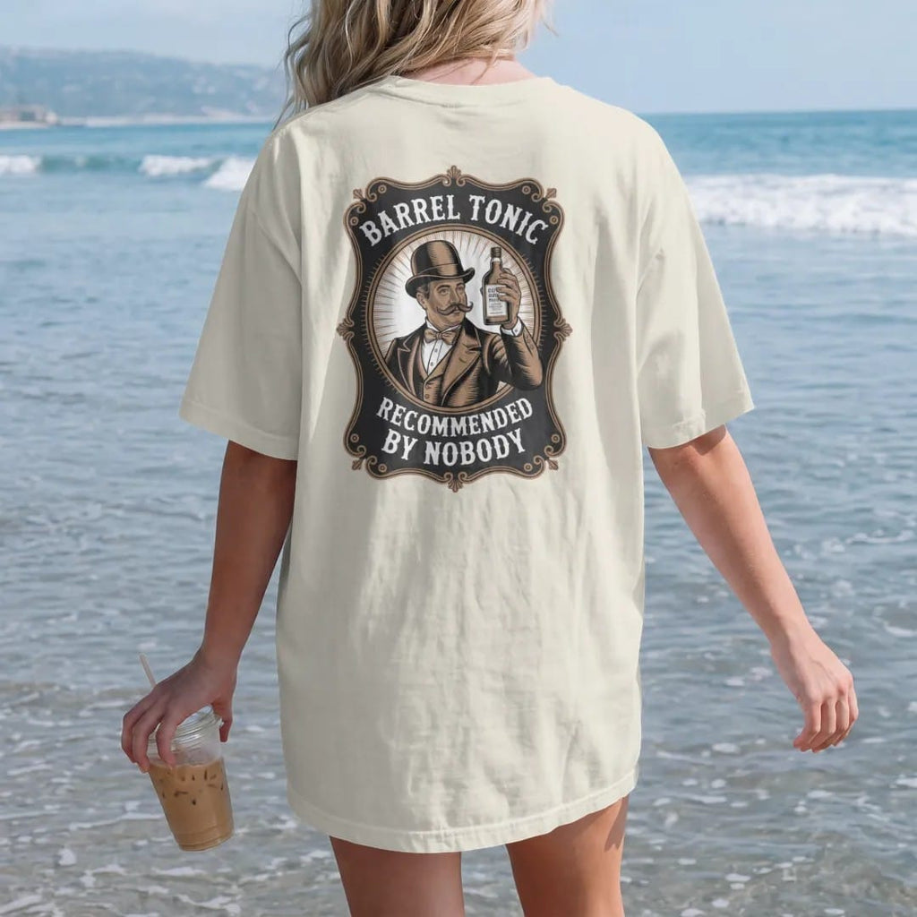 Woman on the beach in a Ivory tee, back view: a mustached gentleman in a bowler hat holding up a whiskey bottle, with "BARREL TONIC" and "RECOMMENDED BY NOBODY", printed as a fine engraving in antique gold and warm cream.