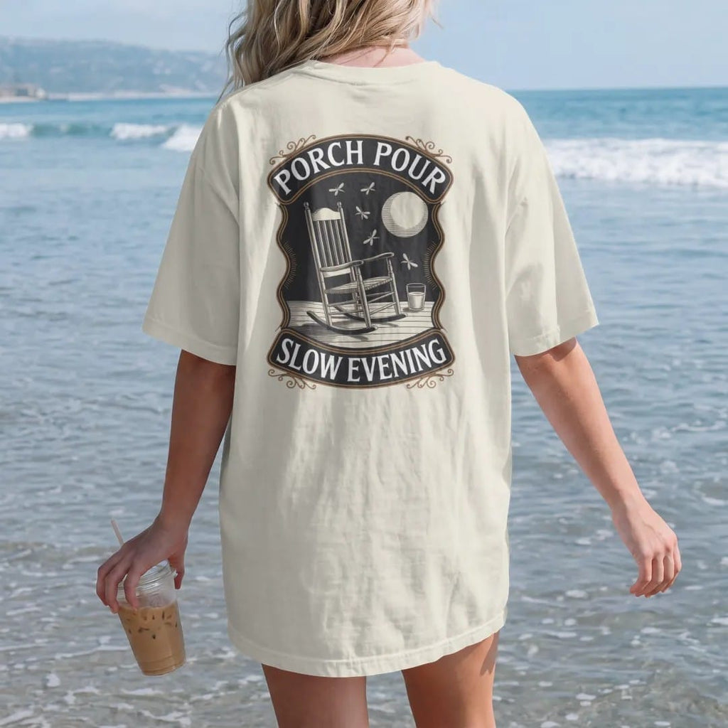 Woman on the beach in a Ivory tee, back view: a rocking chair on a porch with a glass on the armrest under a low moon, with "PORCH POUR" and "SLOW EVENING" in warm Victorian serif capitals, printed as a fine engraving in antique gold and warm cream.