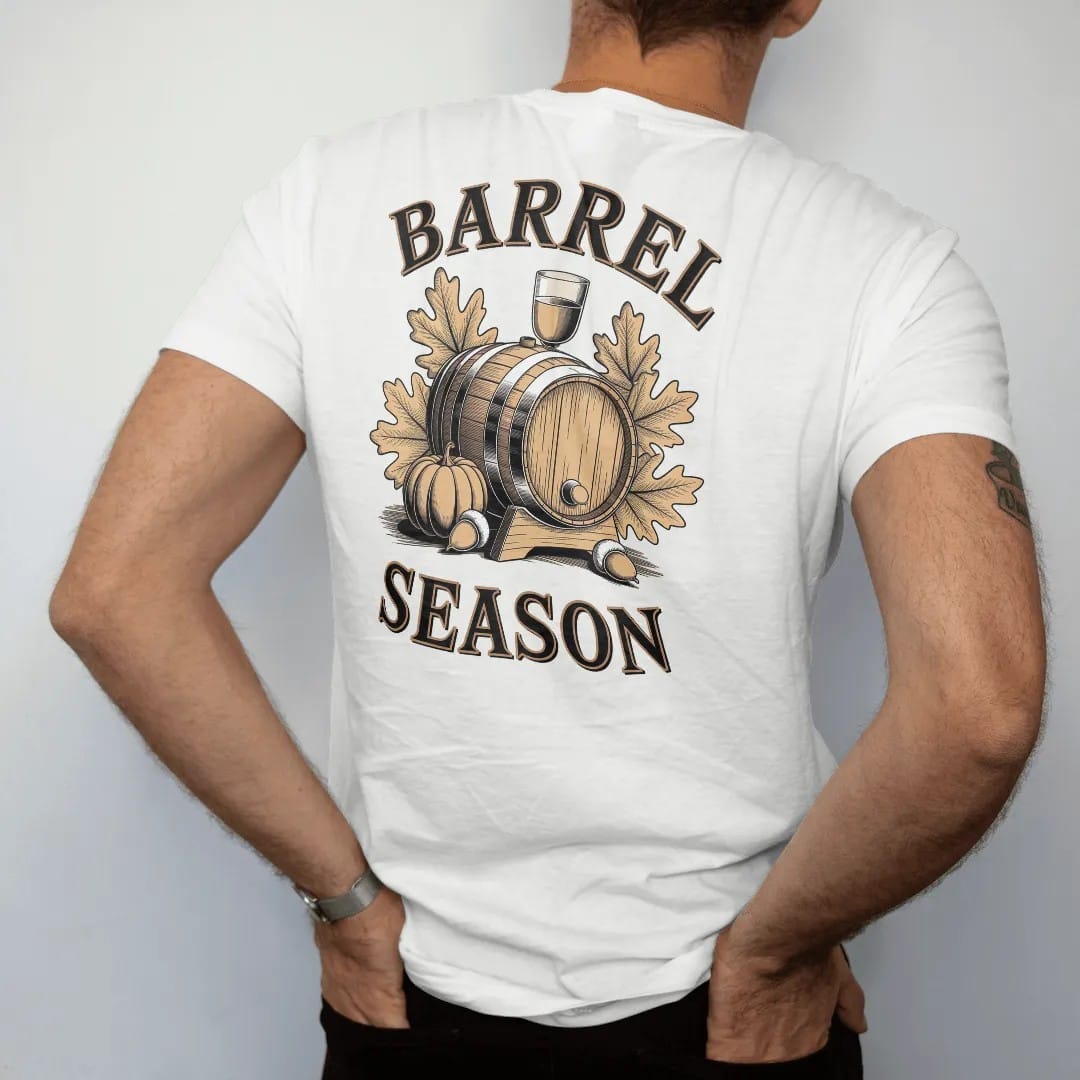Man wearing a White tee, seen from the back: an oak barrel with autumn leaves, acorns and a pumpkin, with "BARREL SEASON" in warm Victorian serif capitals, printed as a fine engraving in antique gold and warm cream.