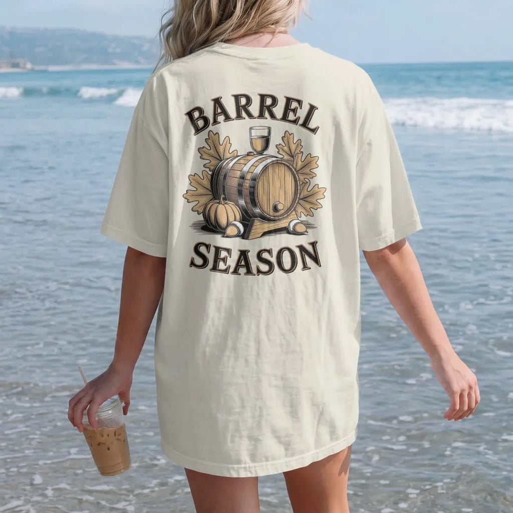 Woman on the beach in a Ivory tee, back view: an oak barrel with autumn leaves, acorns and a pumpkin, with "BARREL SEASON" in warm Victorian serif capitals, printed as a fine engraving in antique gold and warm cream.