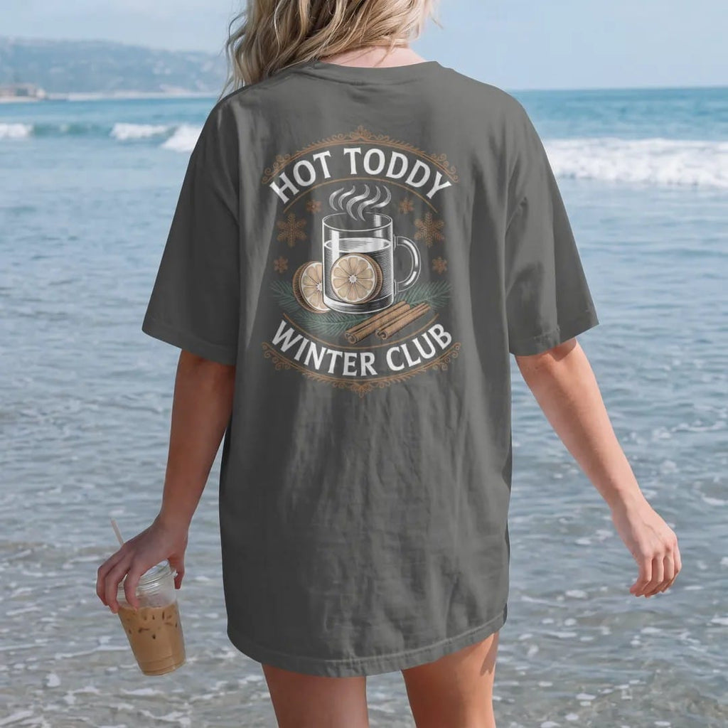 Woman on the beach in a Pepper tee, back view: a steaming toddy mug with lemon and cinnamon among snowflakes, with "HOT TODDY" and "WINTER CLUB" in elegant Didone capitals, printed as a fine engraving in antique gold and warm cream.