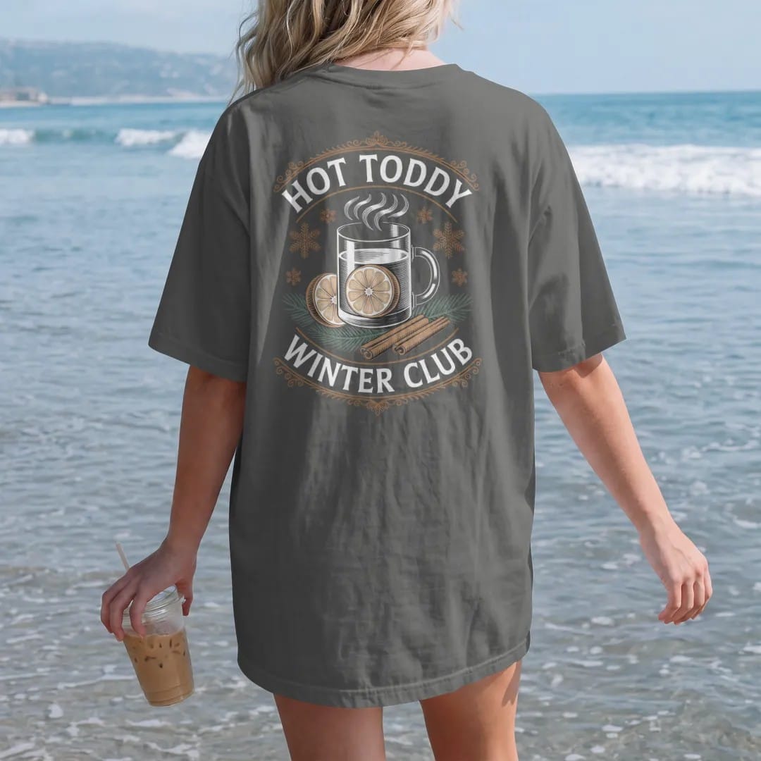 Woman on the beach in a Pepper tee, back view: a steaming toddy mug with lemon and cinnamon among snowflakes, with "HOT TODDY" and "WINTER CLUB" in elegant Didone capitals, printed as a fine engraving in antique gold and warm cream.