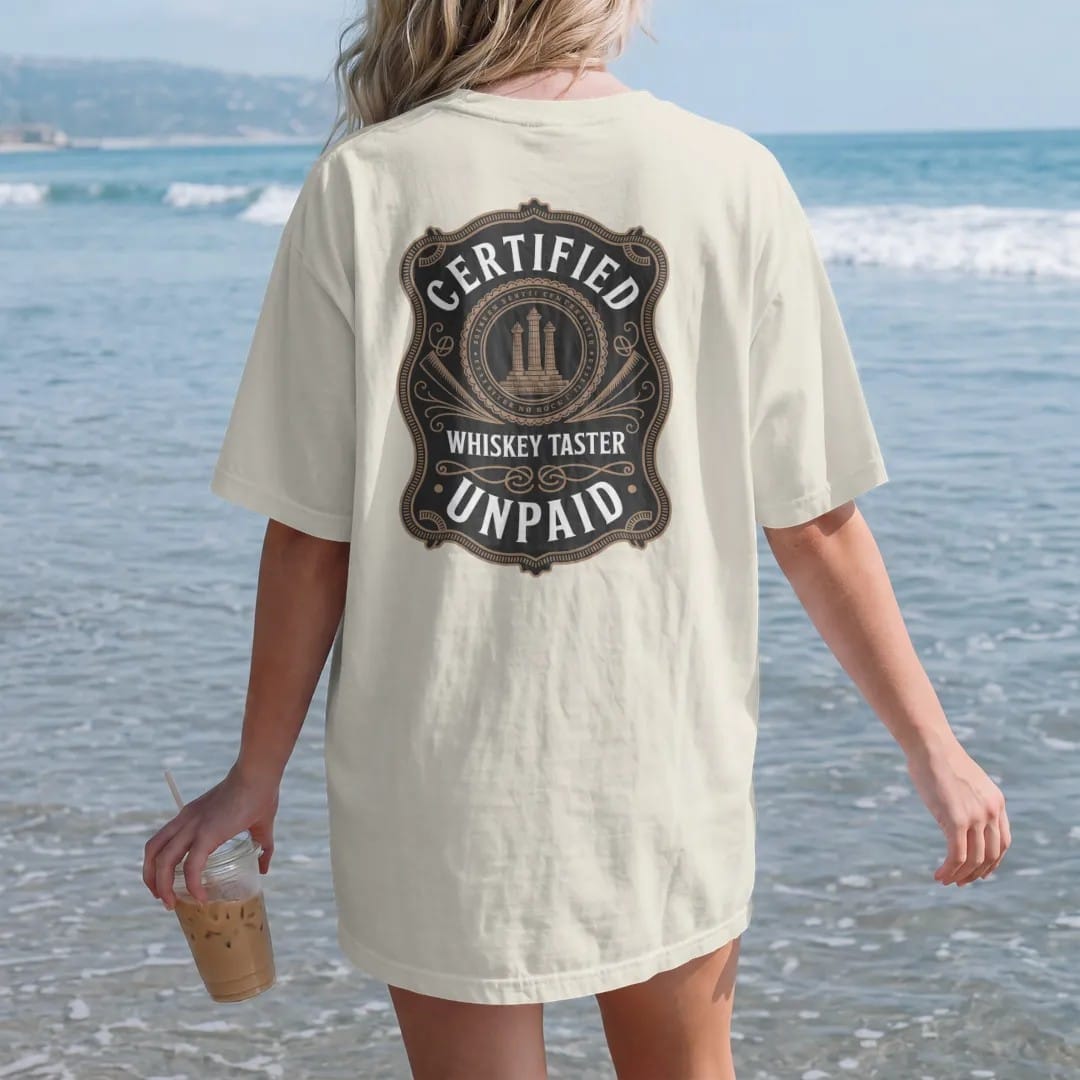 Woman on the beach in a Ivory tee, back view: a diploma-style seal with a glencairn glass and ribbon tails, with "CERTIFIED", "WHISKEY TASTER" and "UNPAID", printed as a fine engraving in antique gold and warm cream.