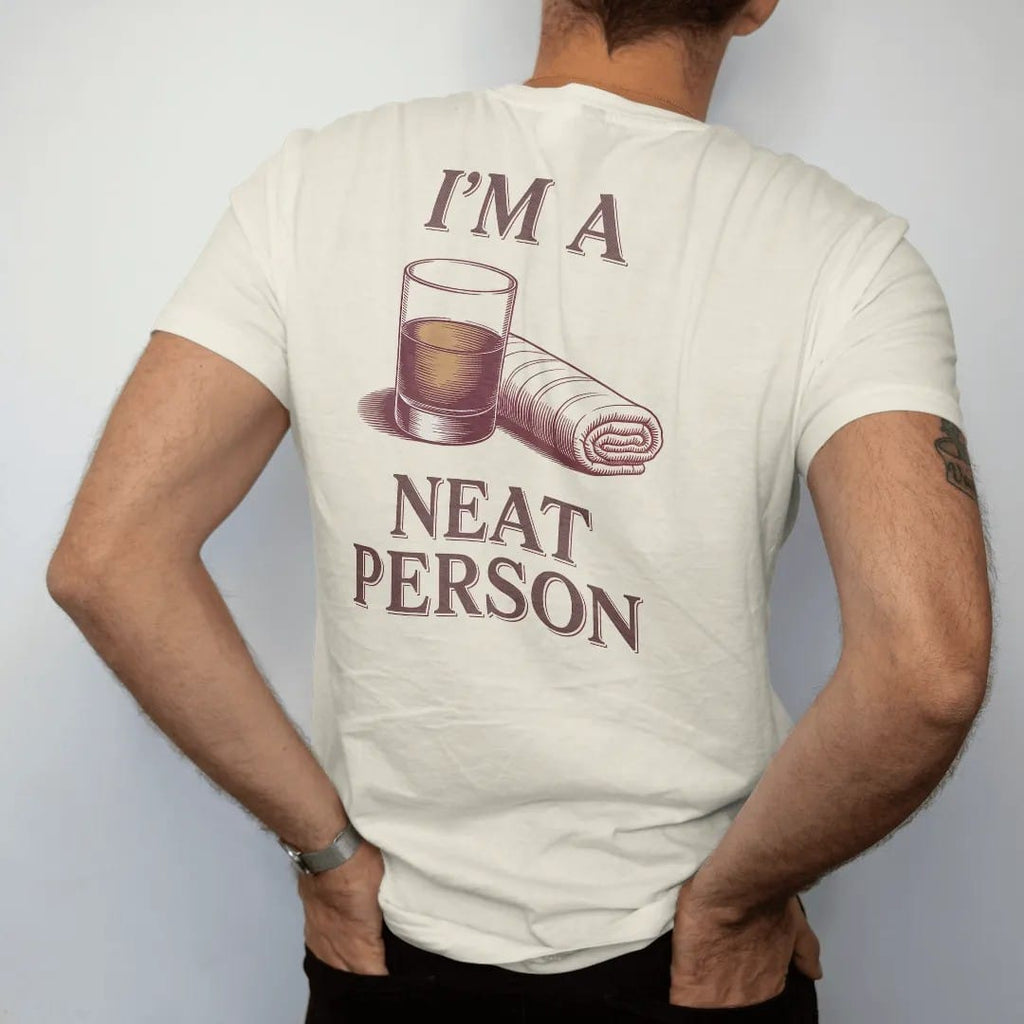 Man wearing a Ivory tee, seen from the back: a rocks glass with a short pour beside a folded bar towel, with "I'M A" and "NEAT PERSON" in elegant Didone capitals with small script, printed as a fine engraving in oxblood, charred oak brown and…