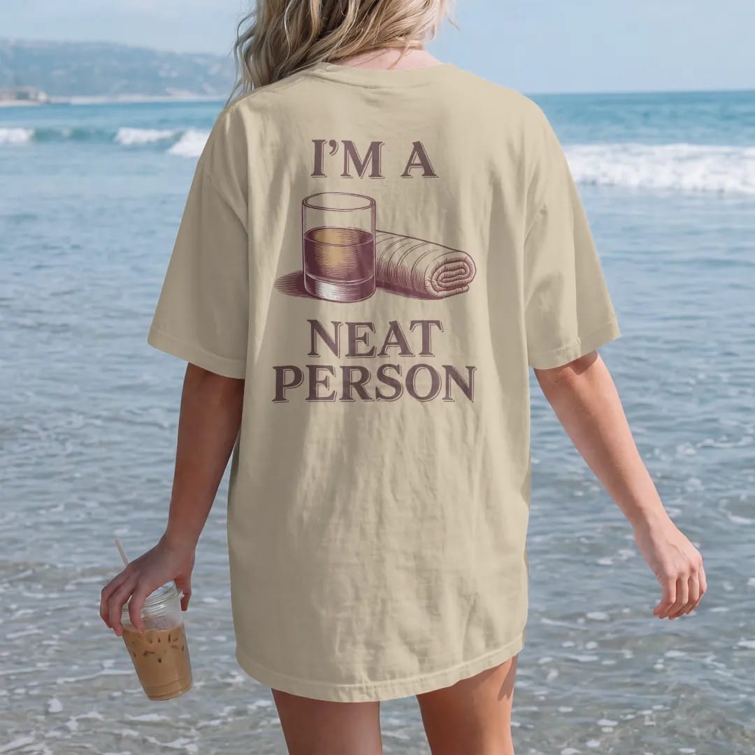 Woman on the beach in a Sandstone tee, back view: a rocks glass with a short pour beside a folded bar towel, with "I'M A" and "NEAT PERSON" in elegant Didone capitals with small script, printed as a fine engraving in oxblood, charred oak brown and…