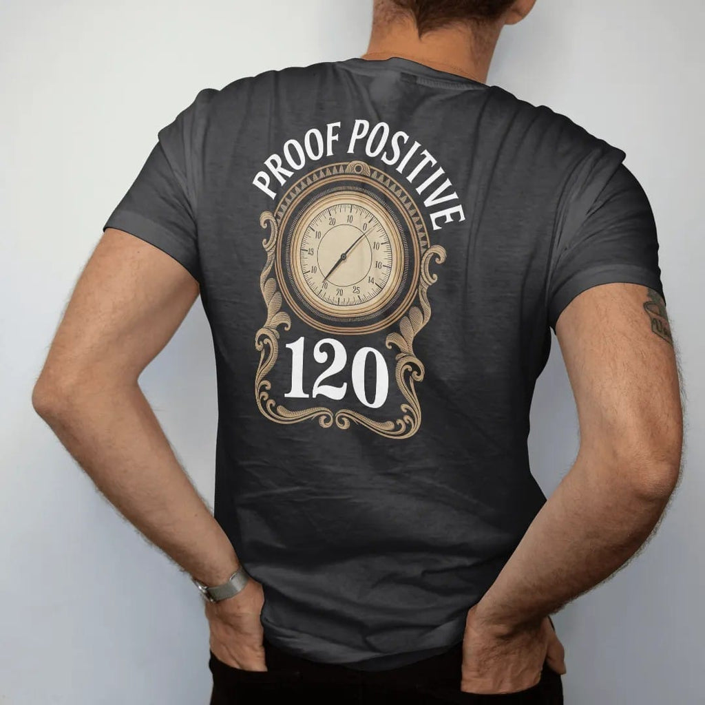 Man wearing a Black tee, seen from the back: an ornate numeral inside an engraved gauge dial, with "PROOF POSITIVE" and "120" in bold Didone numerals and engraved small caps, printed as a fine engraving in antique gold and warm cream.