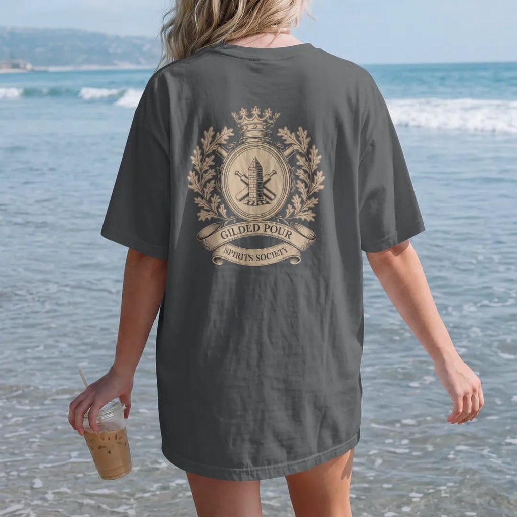Woman on the beach in a Graphite tee, back view: a heraldic crest with a glencairn, crossed bottles and oak-leaf filigree, with "GILDED POUR" and "SPIRITS SOCIETY" in ornate engraved Didone capitals, printed as a fine engraving in antique gold and…