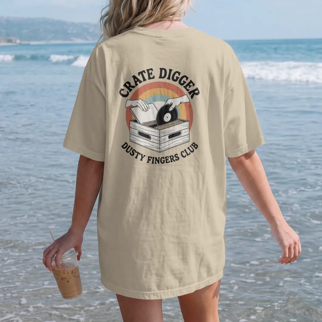 Woman on the beach in a Sandstone tee, back view: two hands flipping through a wooden crate of record sleeves, with "CRATE DIGGER" and "DUSTY FINGERS CLUB" in chunky 70s lettering and small 70s caps, printed in faded, cracked cream, mustard, burnt…