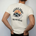 Man wearing a Ivory tee, seen from the back: a turntable tonearm lowering onto a spinning record, with "NEEDLE DROP" and "HOLD YOUR BREATH" in chunky 70s lettering and small 70s caps, printed in faded, cracked cream, mustard, burnt orange and faded…
