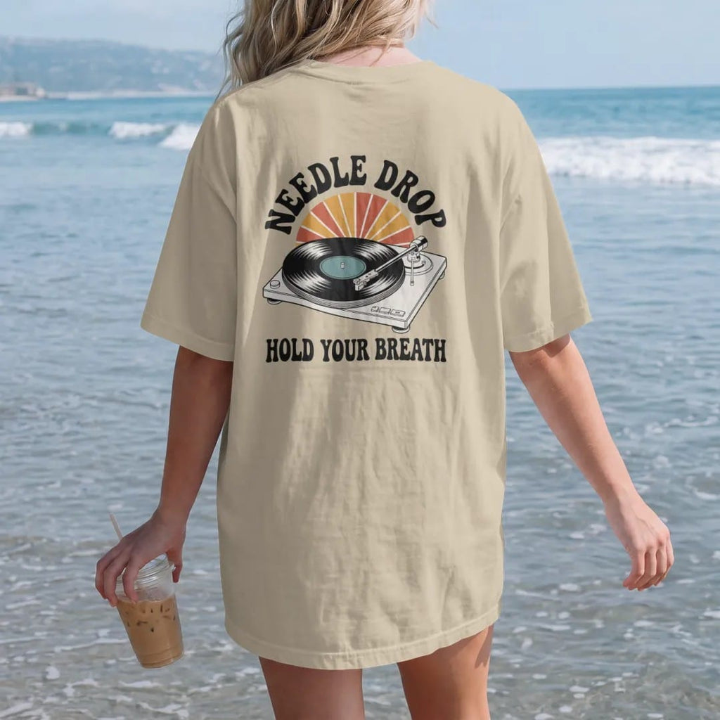 Woman on the beach in a Sandstone tee, back view: a turntable tonearm lowering onto a spinning record, with "NEEDLE DROP" and "HOLD YOUR BREATH" in chunky 70s lettering and small 70s caps, printed in faded, cracked cream, mustard, burnt orange and…